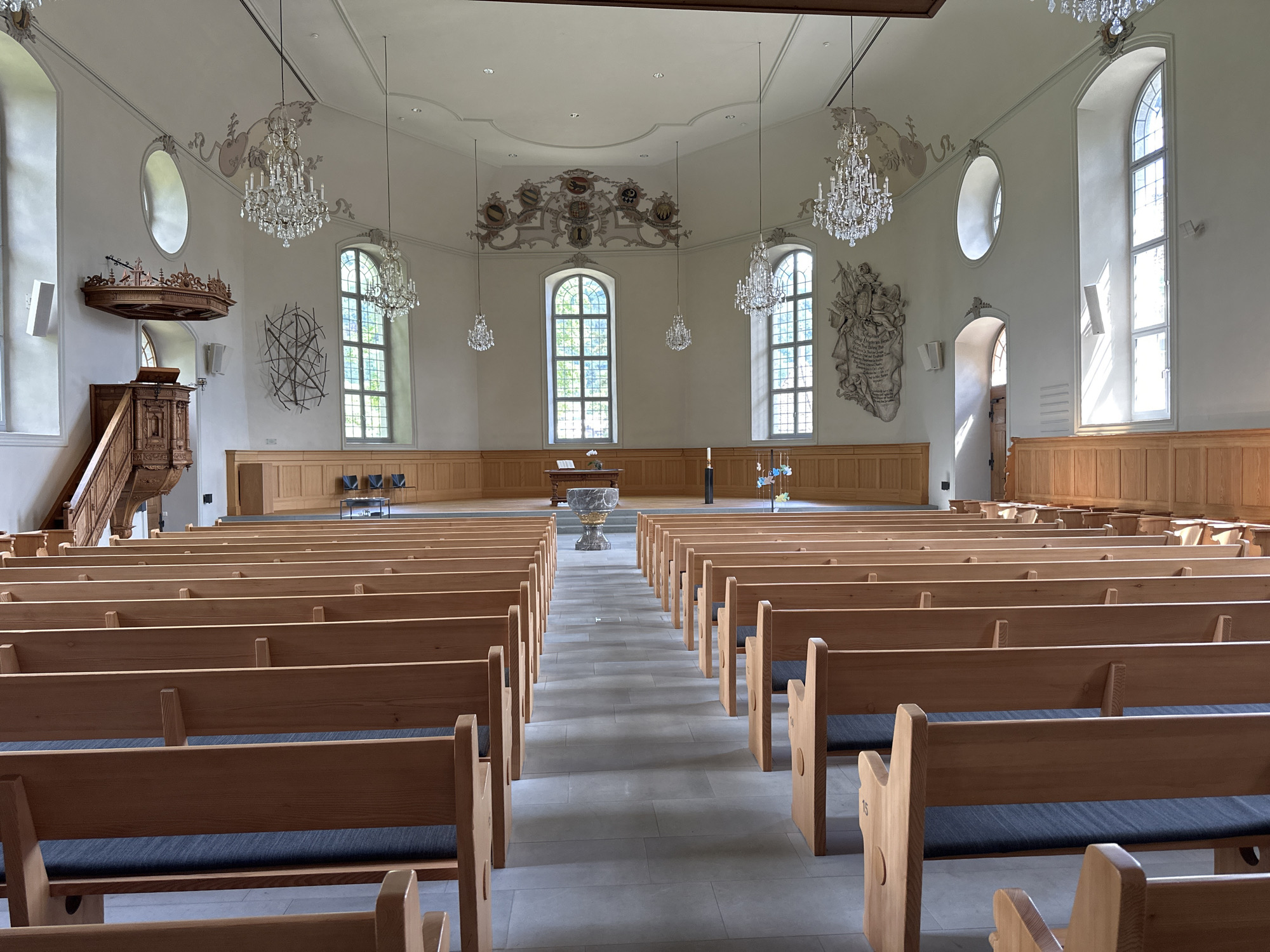 This is a wide-angle interior shot of a bright, airy church in Thun, Switzerland. The scene is empty, with rows of light-colored wooden pews filling the nave. Sunlight streams in through tall, arched windows on the right side of the frame, casting geometric shadows on the floor. Several ornate crystal chandeliers hang from the high, white ceiling, reflecting the light.
The church features traditional elements such as a wooden pulpit on the left, adorned with intricate carvings. Towards the front, an altar with a baptismal font is visible. The walls are decorated with paintings and relief sculptures, including coats of arms and a large, detailed mural on the right. The overall impression is one of solemnity and quietude, with the empty pews suggesting a moment before or after a service. The time of day appears to be daytime, with bright natural light indicating clear weather.