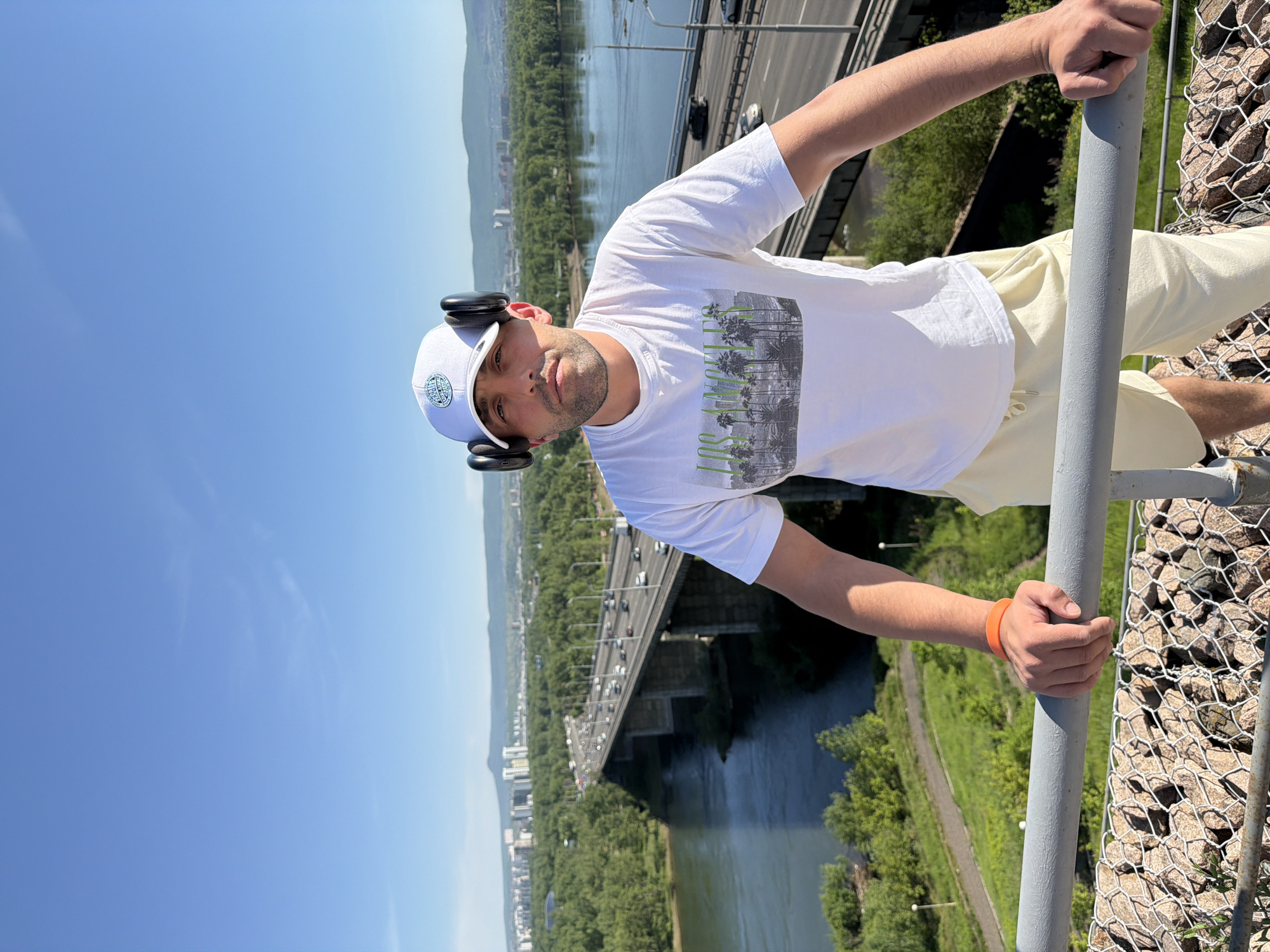 A man stands in an outdoor elevated setting, looking directly at the camera. He wears a white baseball cap with headphones over it, a white T-shirt featuring a "LOS ANGELES" graphic with palm trees, light-colored shorts, and an orange wristband on his left arm. His hands rest on a gray metal railing in the foreground, below which a gabion wall filled with rocks is visible. The background reveals a wide, multi-lane bridge with numerous vehicles spanning a broad river. Lush green trees densely cover the riverbanks and surrounding areas. Beyond the river and foliage, a distant cityscape with various buildings is discernible, backed by mountains under a clear blue sky. This scene is situated in Krasnoyarsk, Russia, specifically at an elevated viewpoint overlooking the **October Bridge** (Октябрьский мост) over the **Yenisei River**.