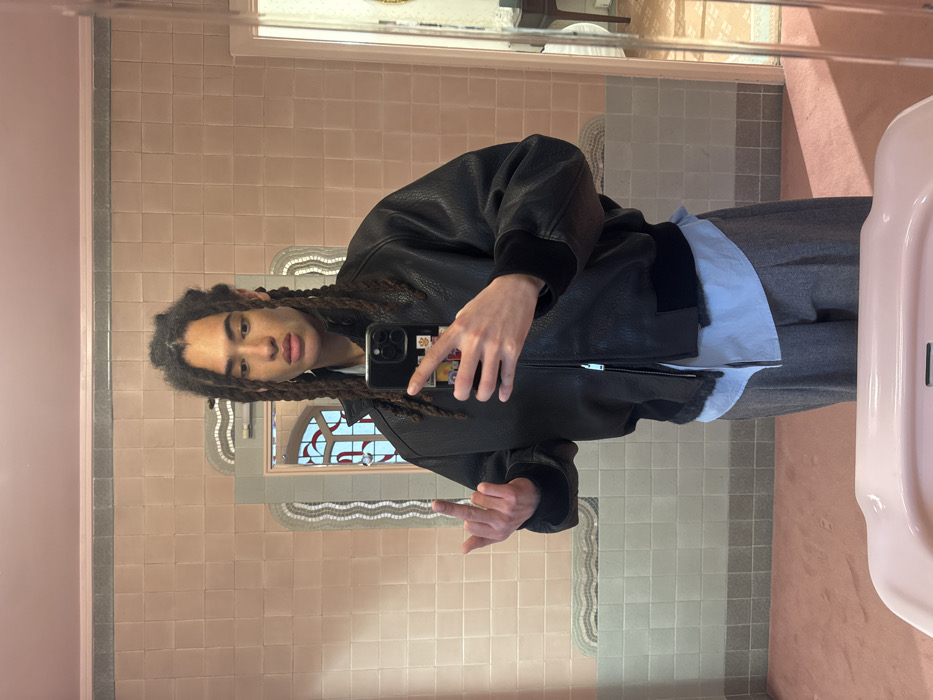An individual with long, dark, braided hair is depicted from the chest up, reflected in a mirror. They are wearing a black bomber jacket with ribbed cuffs and collar, layered over a light blue collared shirt and gray pants. The jacket appears to be made of a textured material, possibly leather or faux leather. The person's left arm is bent at the elbow, and they are holding a dark-colored smartphone, likely an iPhone, in their left hand, which covers part of their face from the viewer's perspective. The phone case has colorful, potentially cartoon-like, stickers or designs visible on its back. The person's right arm is also bent, and their hand is raised slightly, making a gesture that appears to be a "hang loose" or "shaka" sign, with the thumb and pinky extended and the middle fingers curled.
The individual has dark eyes, full lips, and their expression is neutral.
The setting appears to be a bathroom, characterized by the tiled walls and a pink sink visible on the right side of the frame. The walls are covered in light pink square tiles, with a horizontal strip of gray tiles separating them near the top and bottom of the visible section. Below the main tile line, there are more light pink tiles on the lower wall. The floor has a mix of light-colored and gray square tiles. In the background, visible through the mirror's reflection, there's another mirror with an arched top, featuring decorative wavy mosaic patterns around its frame. Through this background mirror, a glimpse of another room or hallway with a table and possibly a chair can be seen. The overall lighting suggests an indoor setting, likely artificial light. The image is captured as a mirror selfie, and the original image is rotated 90 degrees clockwise.