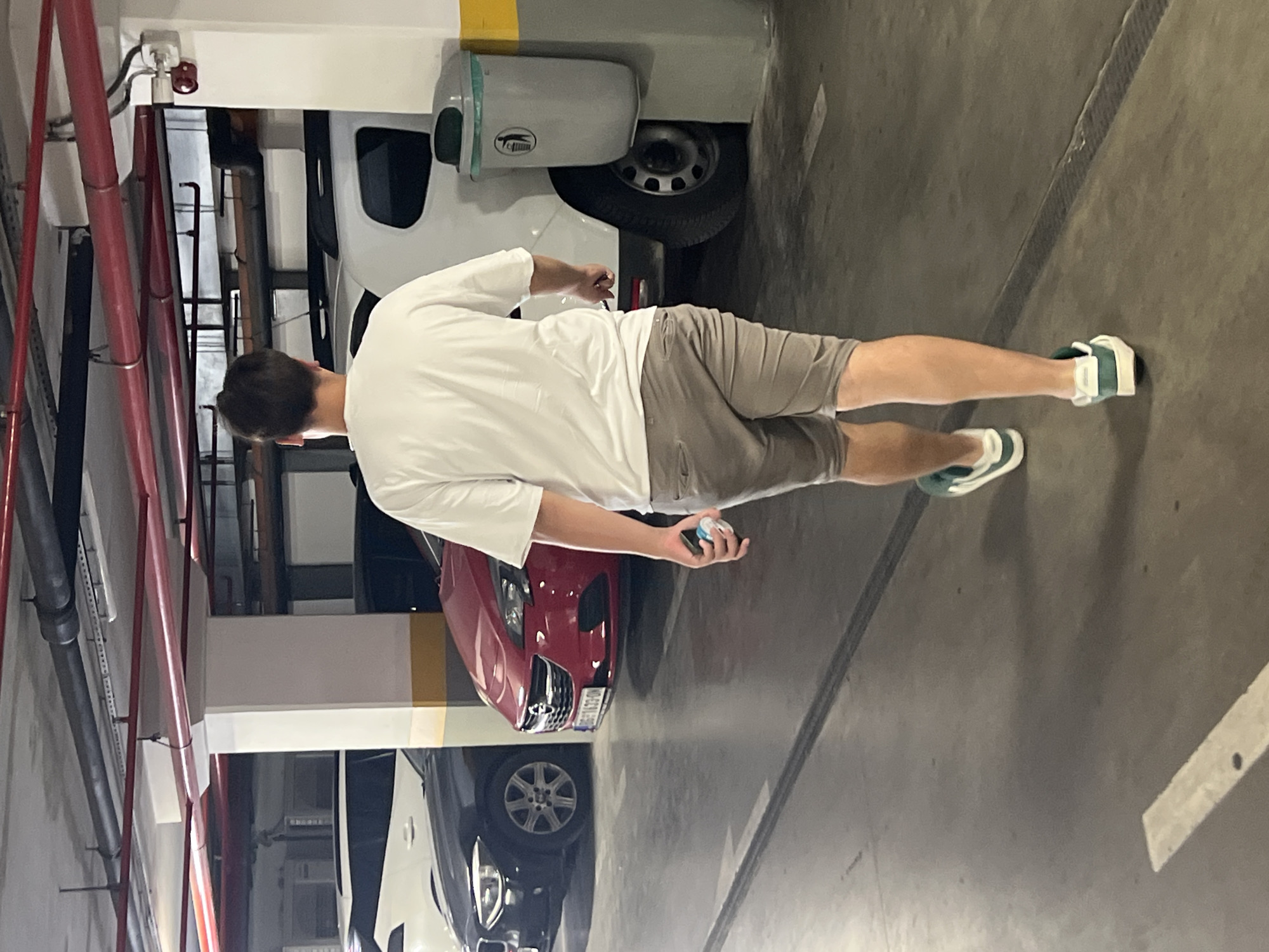 A person is standing in a parking garage in Budva, Montenegro. The person is wearing a white t-shirt and grey shorts, and green and white sneakers. They are holding a small blue and white object in their right hand. The person is facing away from the camera.
The parking garage has concrete floors and walls. There are red pipes running along the ceiling and a white trash receptacle mounted on a light-colored wall. Two cars are parked behind the person: a red car and a dark-colored car. The red car has a license plate with some visible text, but it is not legible. The overall lighting suggests it is daytime, as is typical for a parking garage. The scene is unremarkable, with no indication of any particular event or strong emotion.