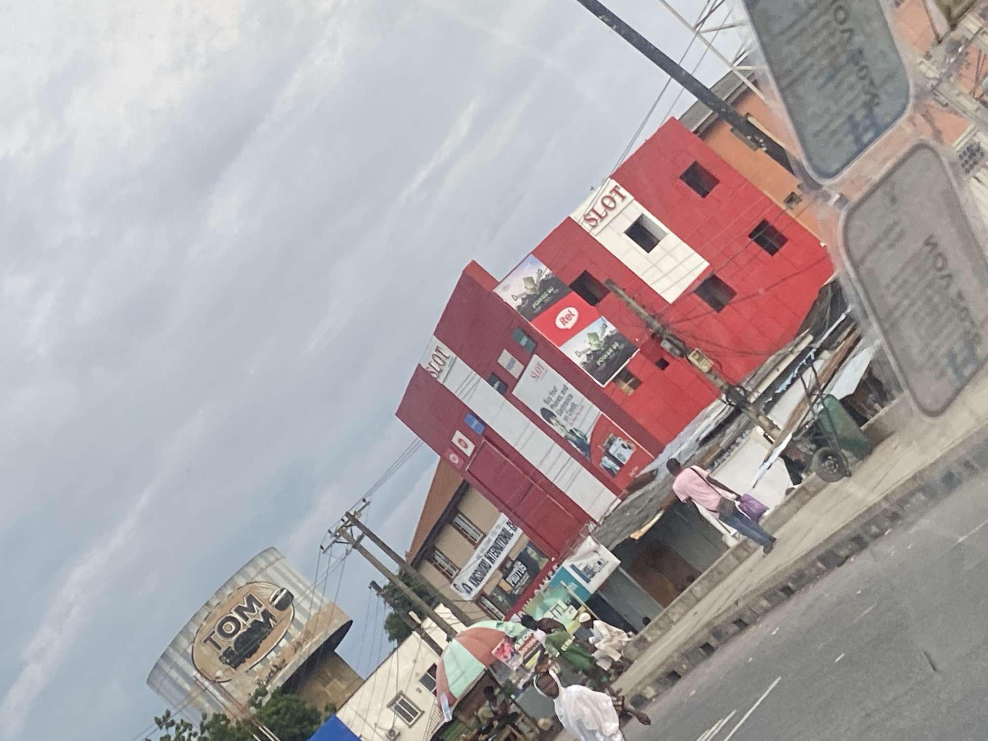 In Ikorodu, Nigeria, a street scene unfolds with a pronounced tilt. A cylindrical billboard with the brand "TOM" is visible against a hazy sky. Dominating the mid-ground is a multi-story red building with white accents, bearing "SLOT" signage. Advertisements for "itel" and "POWER 60" are displayed on its facade. A smaller, cream-colored building with multiple windows stands to the left, featuring a banner that reads "KINGSFORD INTERNATIONAL SCHOOL."
Below these structures, a sidewalk is lined with various stalls and vendors, some with colorful umbrellas. People are seen walking along the sidewalk, including a person in a pink shirt carrying a purple bag. To the left of the sidewalk, a street is visible with a white line marking. Electrical poles and wires crisscross the scene. In the foreground, a blurry reflective surface, possibly the interior of a vehicle, distorts the view of the street and surrounding buildings. The overall impression is of a bustling urban environment under an overcast sky.
