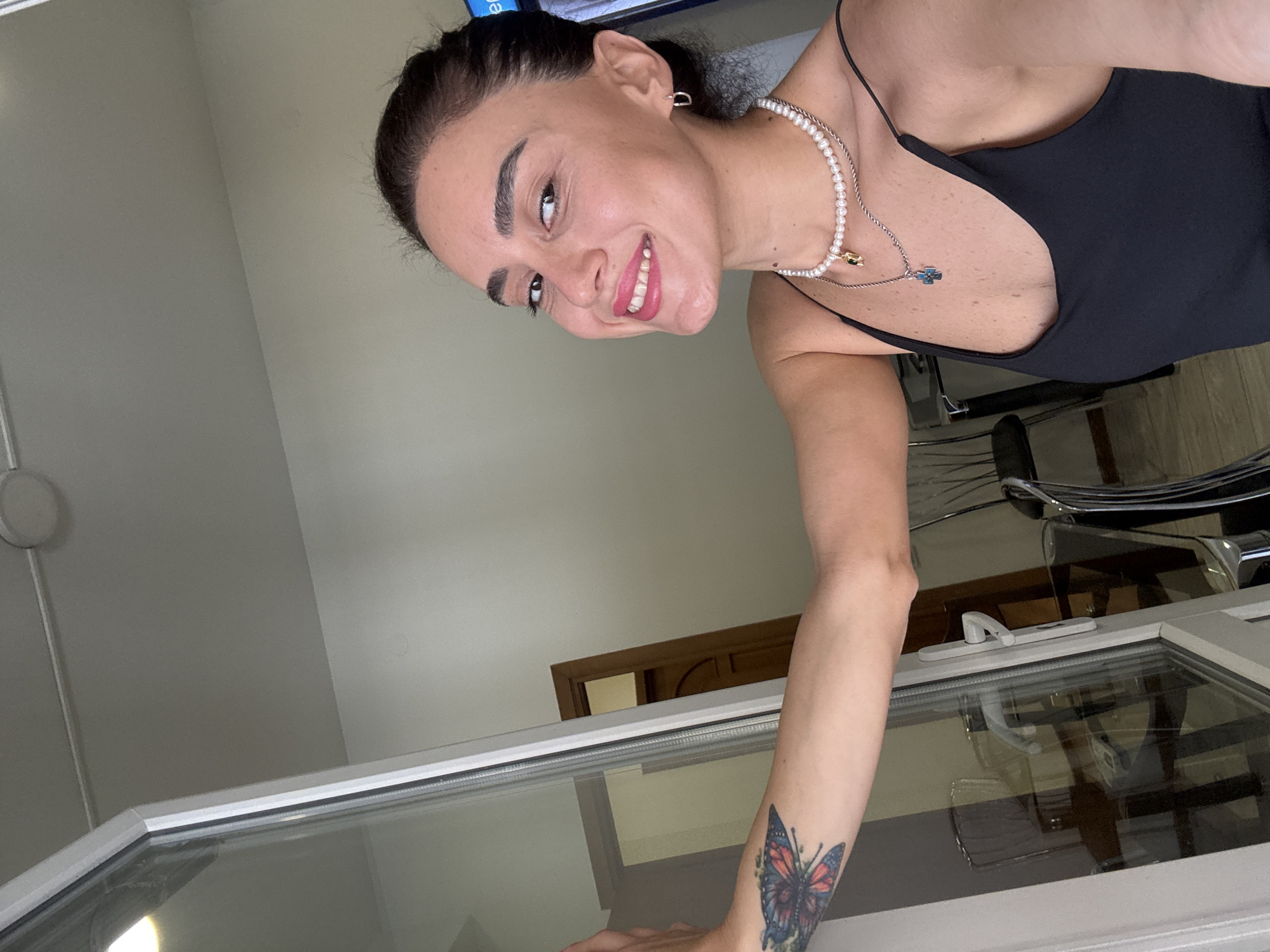 A young woman with dark hair, pulled back into a bun, smiles at the camera. She is wearing a black tank top and two necklaces, one a string of pearls and the other a silver chain with a cross pendant. She has a butterfly tattoo on her left arm. The woman is standing in front of a glass door, which reflects a room with furniture and another door. The background is a light-colored wall with a ceiling light. There are no identifiable landmarks or elements within the image to suggest the specific location of Tbilisi, Georgia.