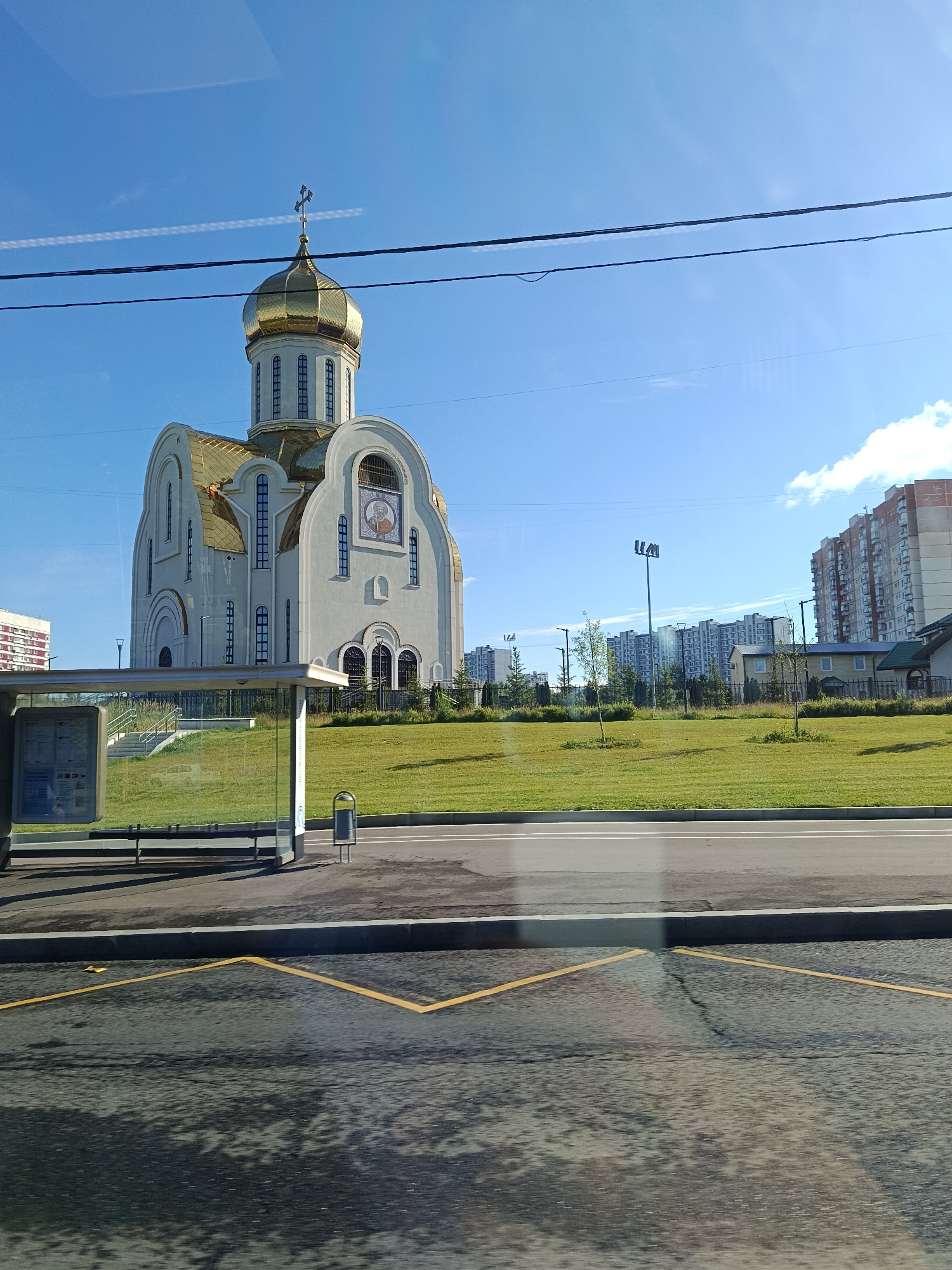 The image shows a contemporary white church with a golden dome and cross, situated on a grassy hill. The church features arched windows and a mosaic icon above the entrance. To the left of the church is a bus stop shelter with a bench and trash receptacle. In the background, a row of multi-story apartment buildings is visible under a clear blue sky with scattered clouds. The foreground displays a street with yellow markings, suggesting a pedestrian crossing or a designated stopping area. The scene appears to be in a suburban or urban residential area.