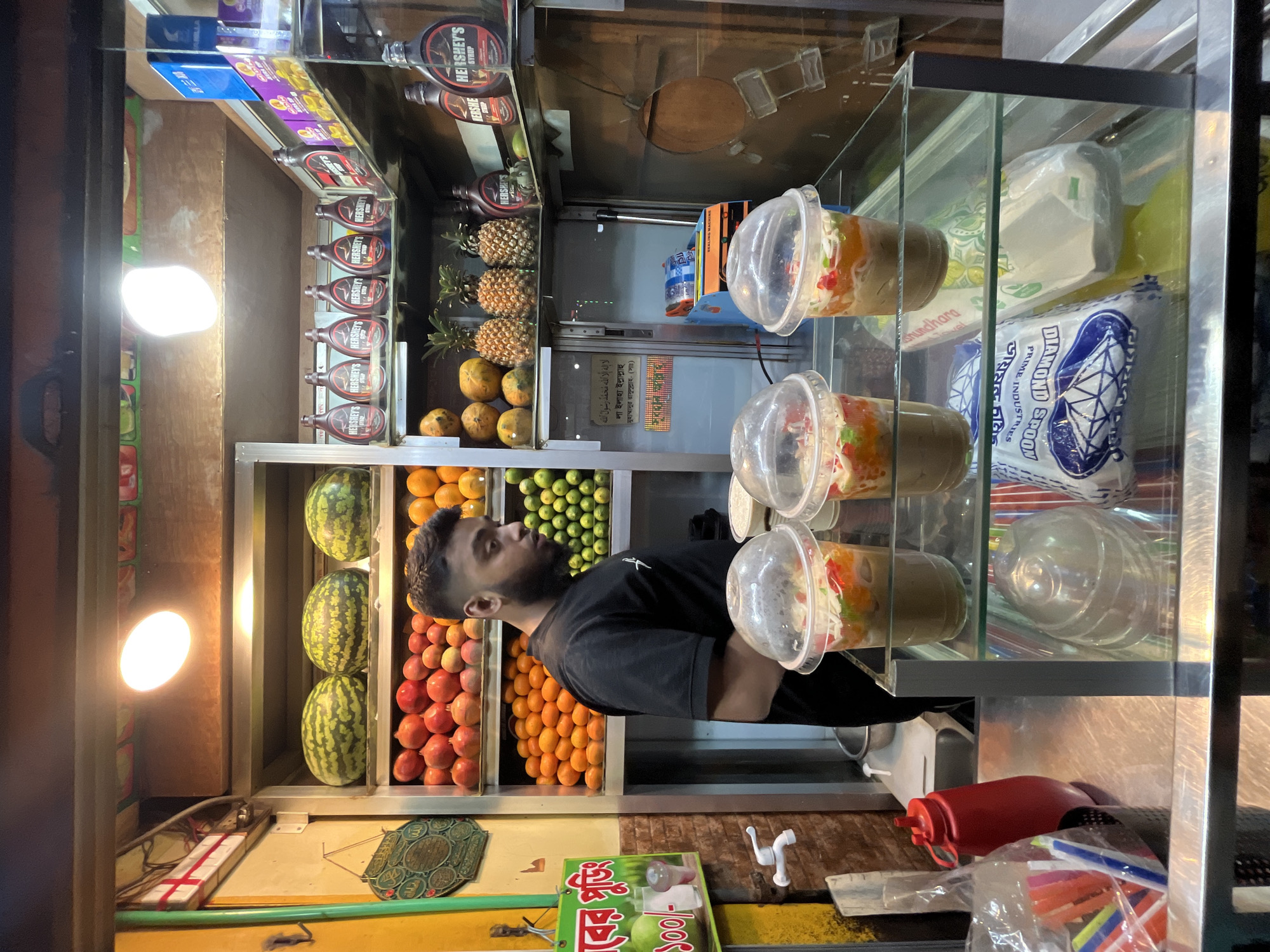 The image captures a street vendor in Dhaka, Bangladesh, likely selling fruit salad or similar refreshments. The scene includes a man, presumably the vendor, standing behind a display of fruits like watermelons, oranges, pomegranates, and pineapples. Above these are rows of Hershey's syrup bottles and packages of Diamond Spoons. In front of the vendor are several plastic containers filled with fruit salad. A sign with text in Bengali and the Shahada, a declaration of faith in Islam, is visible near the bottom of the frame.