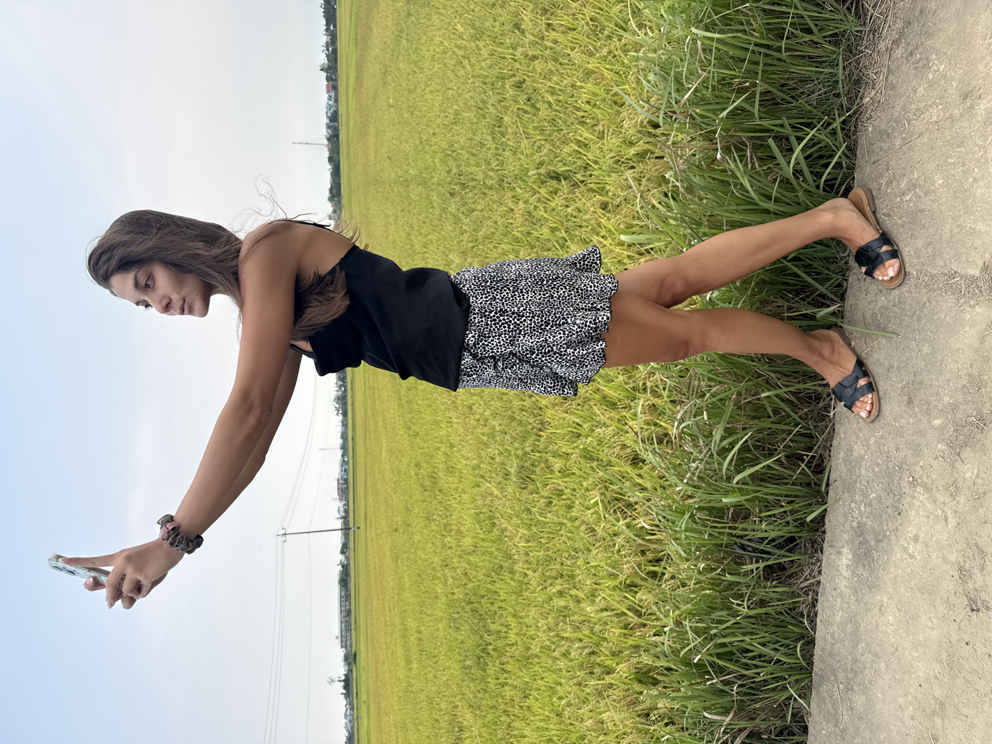 A young woman stands on a narrow concrete path bordering vast agricultural fields, likely rice paddies, stretching into the distance under an overcast sky. She is oriented slightly to her left, extending her left arm forward to hold a light-colored smartphone, possibly engaged in taking a selfie or a photograph. She wears a black tank top, a black and white patterned mini-skirt, and black flat sandals. Her long, brown hair is loose and appears to be gently flowing. A beaded bracelet adorns her left wrist.
The extensive fields behind her are a mix of vibrant green and yellow, indicating a mature crop. Tall green grass grows abundantly along the edge of the concrete path, separating it from the cultivated land. In the background, against the muted sky, faint outlines of distant buildings or structures are visible, along with utility poles and power lines, suggesting a semi-rural setting. The light is soft and diffused, characteristic of an overcast daytime, likely late morning or afternoon. The overall scene evokes a peaceful, rural landscape, consistent with agricultural regions often found near Đà Nẵng, Vietnam. No visible text is present in the image.
