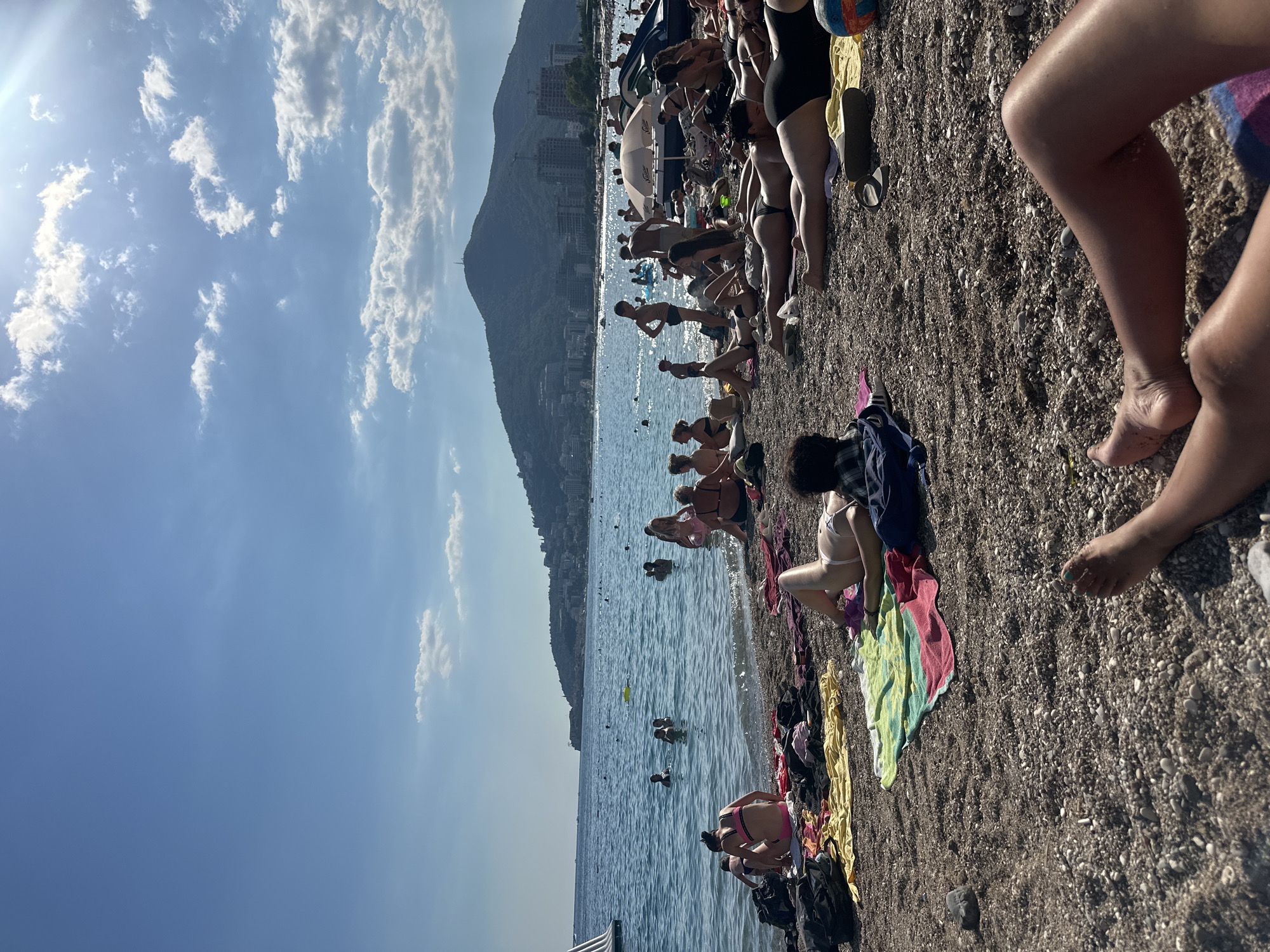 The image depicts a bustling beach scene on a sunny day, likely in Budva, Montenegro, given the characteristic mountainous backdrop and coastal architecture visible in the distance. The foreground is dominated by a pebbly beach strewn with colorful towels, bags, and beach accessories, where numerous individuals are sunbathing and relaxing. Several people are lying on towels, some in swimwear, others with clothing partially on. In the middle ground, the clear blue sea is populated by many people swimming and wading. The water is calm with gentle waves lapping at the shore. Beyond the swimmers, a variety of watercraft, including a jet ski and what appears to be a small sailboat or windsurfer, are present. The background features a large, verdant mountain with buildings clustered along its slopes and at its base, suggesting a coastal town. The sky is a bright blue with scattered white clouds, and the sun casts a strong light, creating bright highlights and some shadows on the beach. The overall atmosphere is one of leisure and summer recreation.