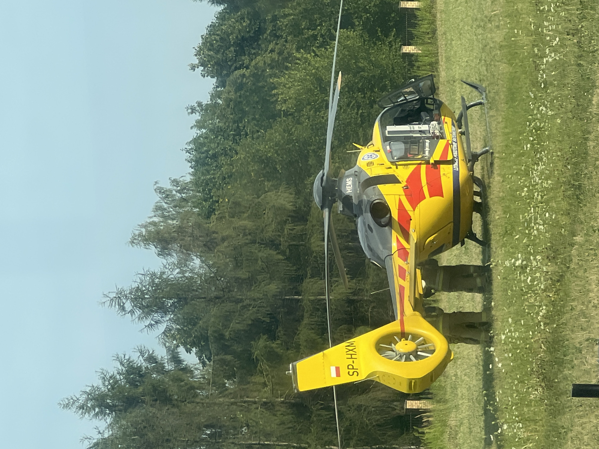 A yellow and red HEMS (Helicopter Emergency Medical Service) helicopter, with registration SP-HXM, is parked on a grassy field. Two individuals in dark uniforms, possibly emergency responders, stand near the rear of the helicopter. The helicopter has its main rotor blades extended and the rear rotor is visible. The background consists of a dense line of green trees and a clear sky. A low brick fence is partially visible to the right of the helicopter. The scene appears to be in an open area, possibly on the outskirts of Lublin, Poland.