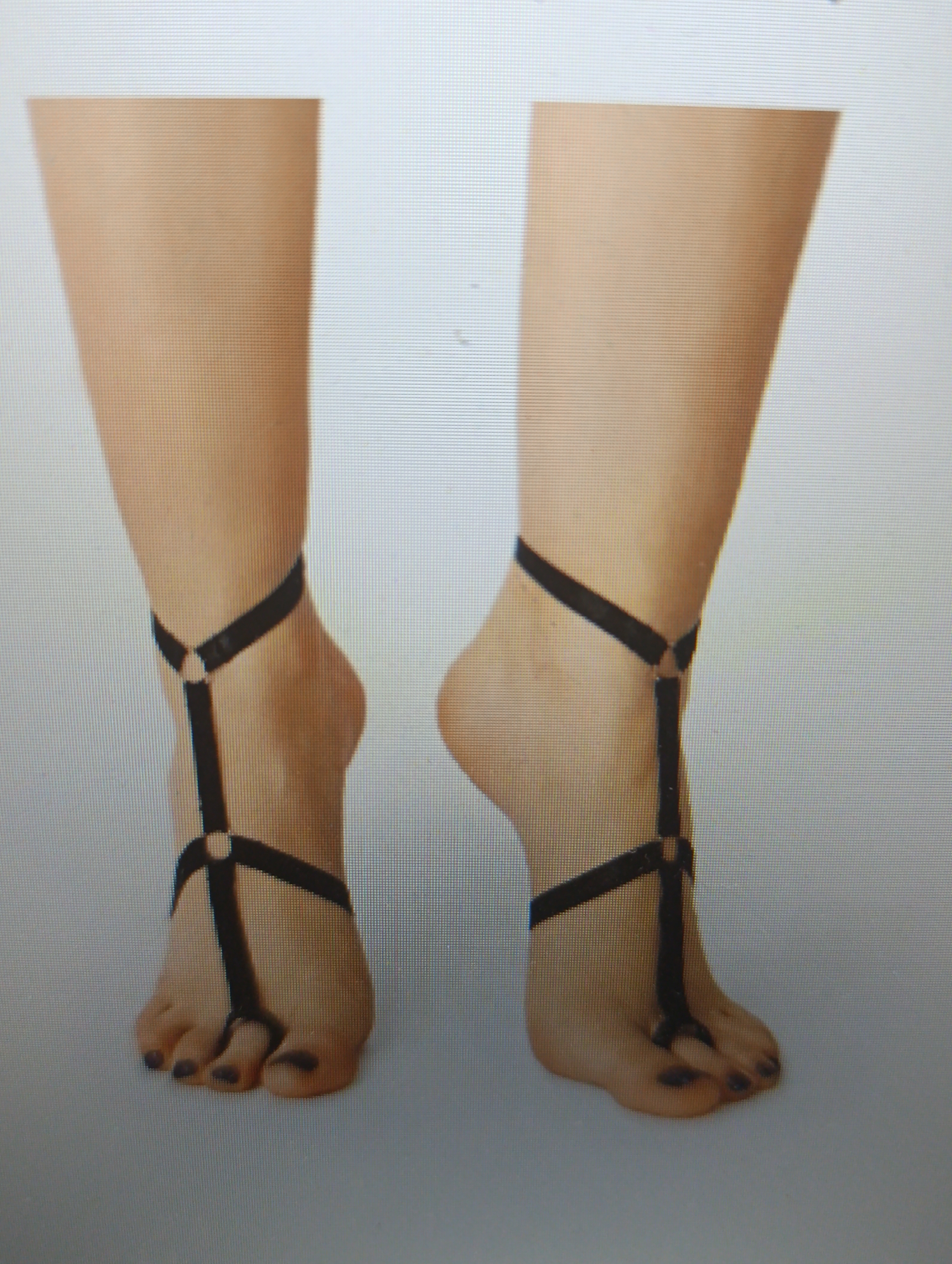 Two bare human feet, adorned with black garter-like straps that wrap around the ankles and extend to the toes, are presented in a close-up, head-on view. The skin tone is light, and the toenails are painted a dark color. The background is plain and light-colored, suggesting a studio or isolated setting. The lighting is even, highlighting the details of the feet and the straps.