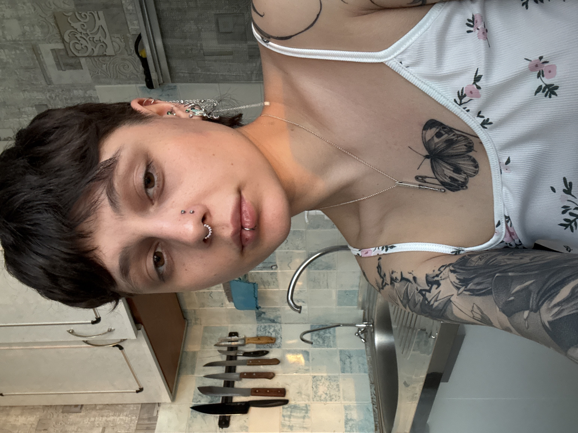A young individual is featured in a close-up, rotated portrait, seemingly a selfie taken in a kitchen setting in Saint Petersburg, Russia. The person has short, dark, textured hair and an array of facial and ear piercings, including a septum ring, double nostril piercings, a philtrum piercing, and multiple ear piercings with intricate chains and various jewelry. Their expression is neutral, and they look directly at the camera with brown eyes.
They are wearing a white ribbed tank top adorned with small pink floral patterns. Visible tattoos include a detailed black and white butterfly on the upper chest, and an elaborate, possibly full-sleeve, black and grey tattoo on the right arm, featuring what appears to be a large figure or scene. Another tattoo consisting of black lines is partially visible on the left shoulder/upper arm. A thin silver chain necklace with a rectangular pendant hangs around their neck.
The background reveals a kitchen with light blue and off-white patterned tiles on the walls. A set of kitchen knives is neatly arranged on a magnetic strip attached to the tiled wall. A metal kitchen faucet and part of a sink are also visible, along with light-colored kitchen cabinets featuring metal handles. A small blue cloth or sponge is present near the faucet. Natural light suggests it is daytime. There is no visible text in the image.