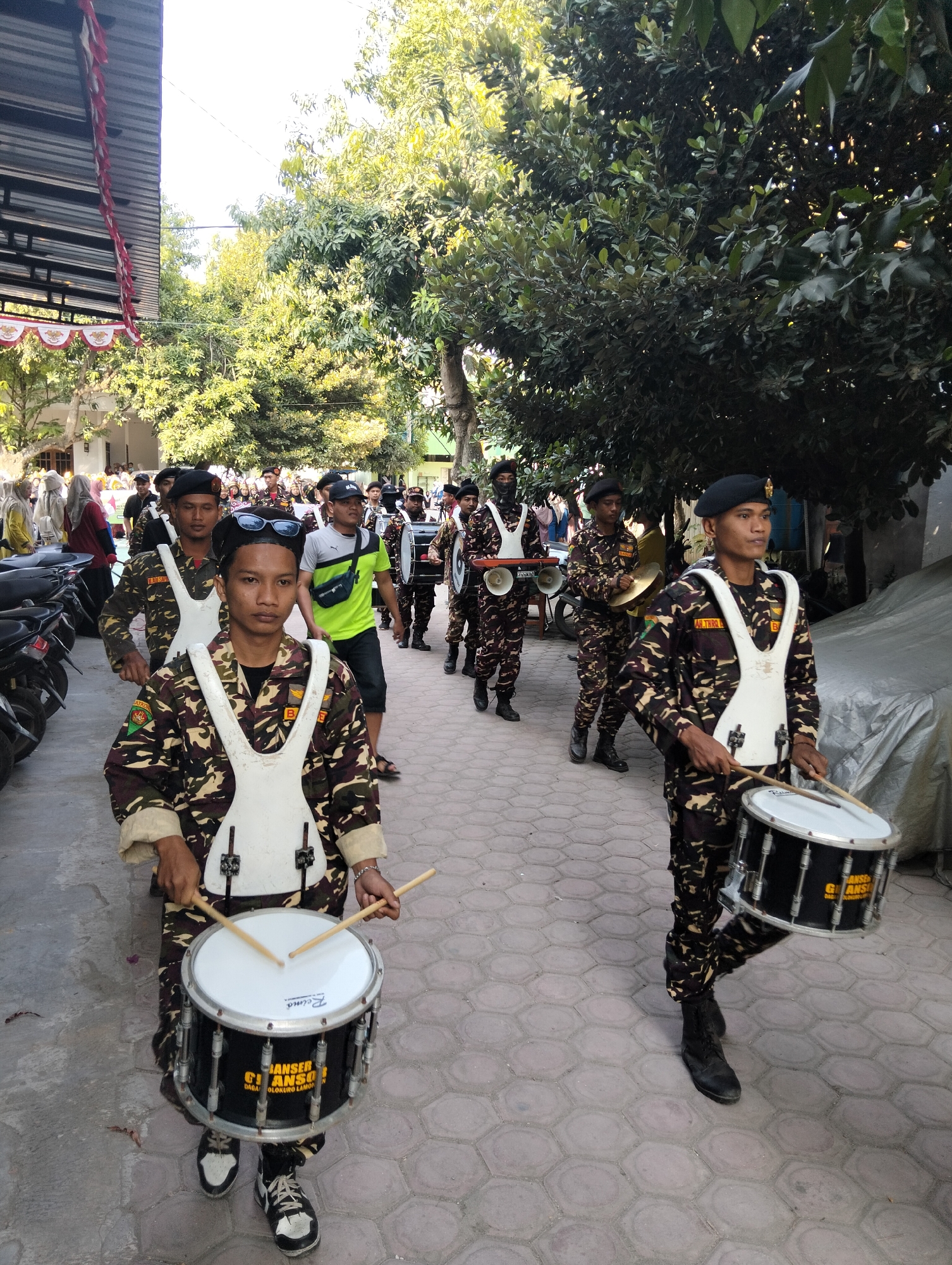 In Kranji, Indonesia, a group of young men in camouflage uniforms march in procession. The uniforms consist of camouflage trousers and jackets with a white vest over them. Some of the individuals are playing musical instruments, including drums and cymbals, as part of what appears to be a marching band. In the foreground, a young man with sunglasses on his head is playing a drum, with drumsticks in his hands. The procession is taking place outdoors on a paved street, with trees and buildings visible in the background. The scene suggests a local event or parade.
