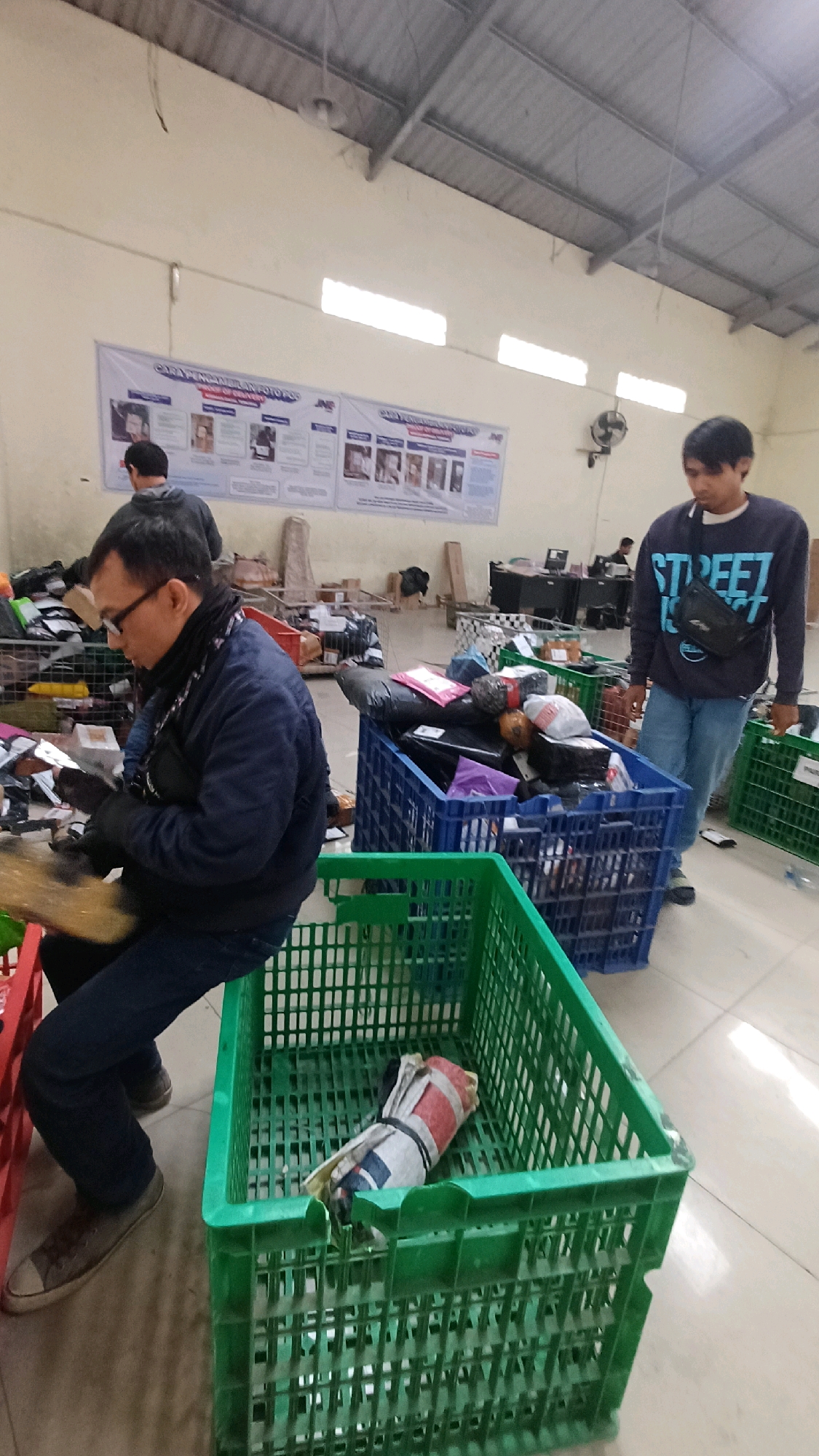 The image shows an interior scene in a warehouse environment, possibly in Kedu, Indonesia. Two men are present. In the foreground, a man wearing a dark jacket, glasses, and a scarf is seated and appears to be sorting through items. He is wearing black gloves and holding a piece of material that is golden-brown in color. To his right, a large green plastic crate contains a rolled-up item that is red, white, and blue. Behind him, another man wearing a dark blue sweatshirt with the word "STREET" on it and blue jeans stands near a blue plastic crate overflowing with packages. The blue crate also contains various smaller packages and bags. In the background, a banner hangs on the wall with images and text. Several other crates and boxes are visible, along with office desks and computers in the far background, suggesting this is a logistics or sorting facility. The floor is tiled. The lighting appears to be from overhead fluorescent fixtures and possibly natural light from high windows.
