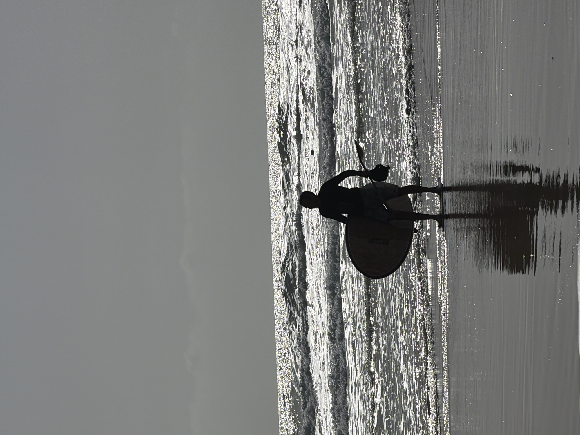 The image depicts a silhouette of a person holding a round object standing on a paddleboard in the water at a beach in Tel Aviv, Israel. The beach is at the water's edge, and small waves reflect the sunlight. The sky is overcast.
