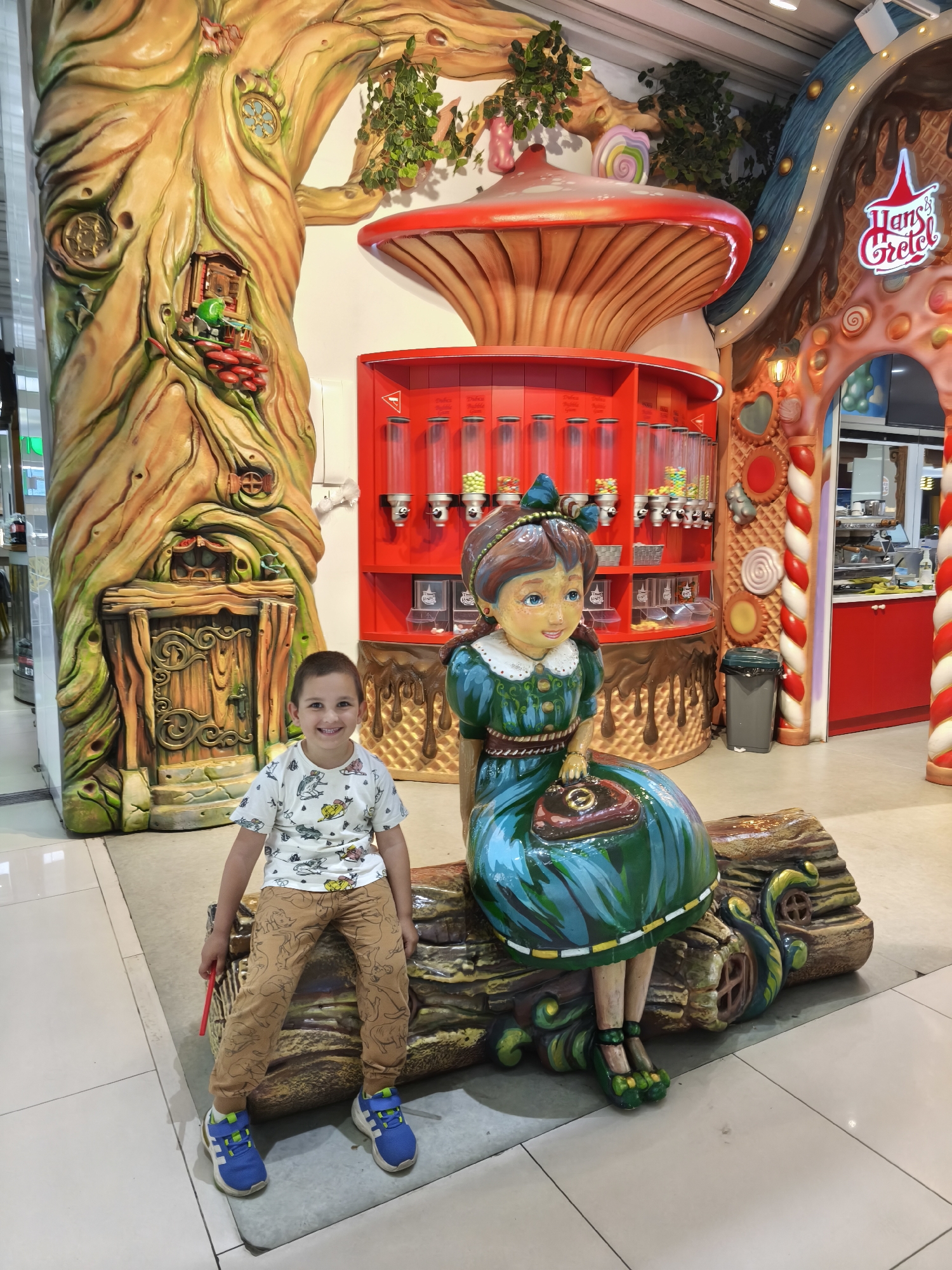 A young boy sits on a log beside a large, painted statue of a seated girl with a purse, located inside a "Hans & Gretel" themed candy shop in Plovdiv, Bulgaria. Behind the boy is a tree structure with a small door, and behind the statue is a red candy dispenser structure resembling a mushroom. The candy shop continues into the background with red and white striped columns and additional candy-themed decorations.