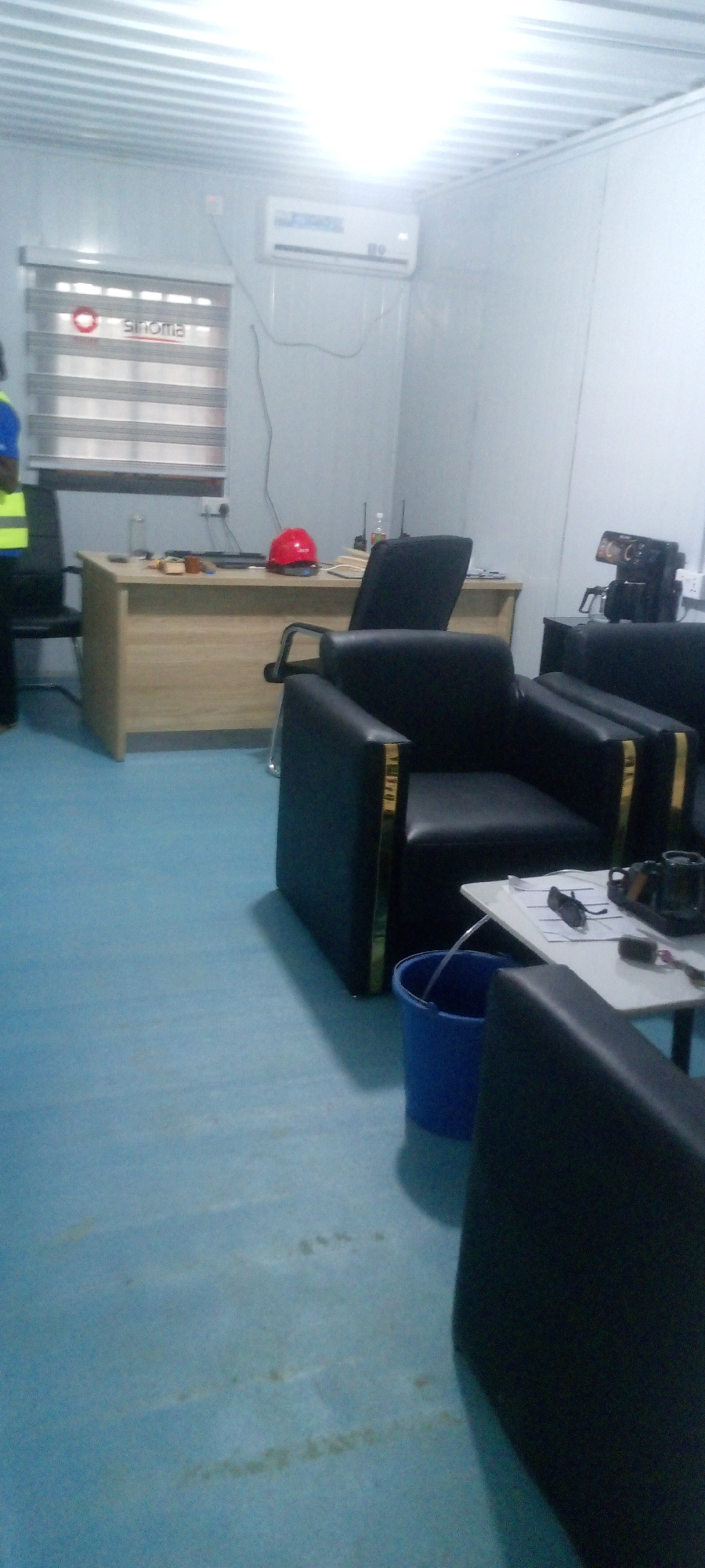 The image depicts an office interior, likely a temporary site office. The room is furnished with a wooden desk, a red hard hat resting on it, and two black leather armchairs with gold trim. A small side table with papers, sunglasses, and a coffee mug is positioned near one of the armchairs. A blue bucket is on the floor, with a tube draining into it. In the background, an air conditioning unit is mounted on the wall above a window with blinds. A person wearing a high-visibility vest stands near the desk. The walls are white, and the floor is a light blue. The ceiling appears to be made of corrugated metal panels. The overall impression is of a functional, albeit sparsely decorated, workspace. There is no specific indication of the city of Indo, Nigeria in the image itself.