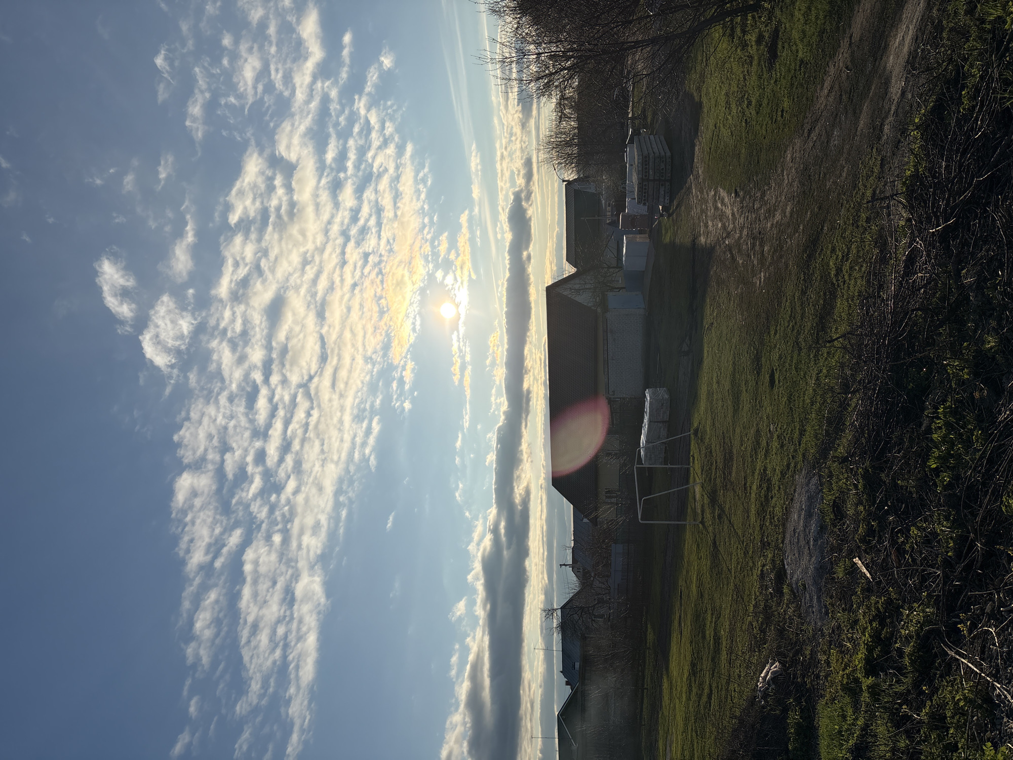 This image, when viewed horizontally, depicts a bright, partly cloudy day in a rural or semi-rural landscape in Ulyanovsk, Russia. The sun, a prominent feature in the upper left, shines intensely, creating a noticeable circular lens flare in the mid-right of the frame. The sky is a clear blue, filled with numerous white, fluffy clouds, many of which are brightly lit by the sun and form distinct bands across the heavens.
The foreground and midground are covered predominantly in green grass, with some visible dirt paths or darker patches of earth. On the right side, a cluster of bare-branched trees and some bushes with emerging green foliage frame the scene. In the midground, several low-rise buildings, likely houses or outbuildings, are scattered, featuring various roof types. Adjacent to some of these structures, stacks of what appear to be construction materials, possibly concrete slabs, are neatly arranged. A white or metallic frame, resembling a small goal or a clothesline, stands in the grassy area. No specific human activity is taking place. There is no visible text within the image. The scene conveys the calm ambiance of a sunny day in a village-like environment.