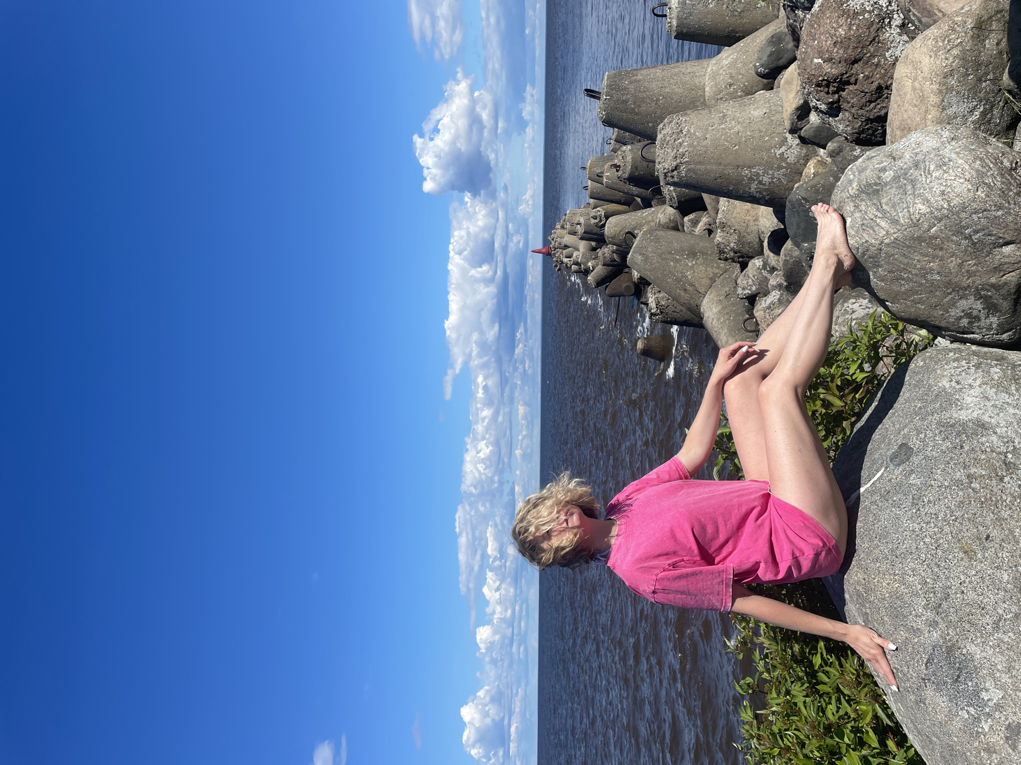 A young woman with blonde hair, wearing a bright pink t-shirt, sits on a large grey rock on the shore of the Beloye Lake in Belozersk, Russia. Her legs are crossed, and she is looking towards the water, with her hair partially obscuring her face. In the background, a long breakwater made of concrete tetrapods extends into the dark blue water under a bright blue sky with fluffy white clouds. The scene is set during the daytime, indicated by the strong sunlight and clear weather.
