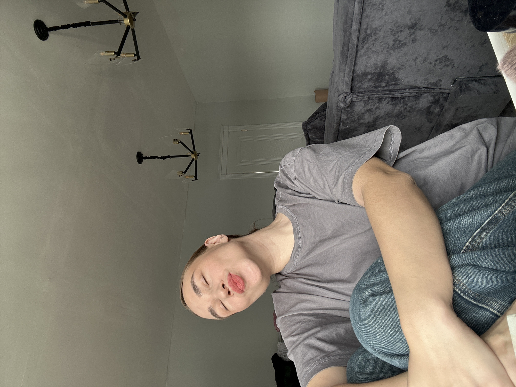 A woman is reclining on a gray upholstered couch indoors. She is wearing a gray t-shirt and blue jeans. Above her are two black and brass chandeliers hanging from a white ceiling. Behind her is a white door. The location is an unidentified indoor setting; there is no information to confirm the city of Kharnavy, Russia.