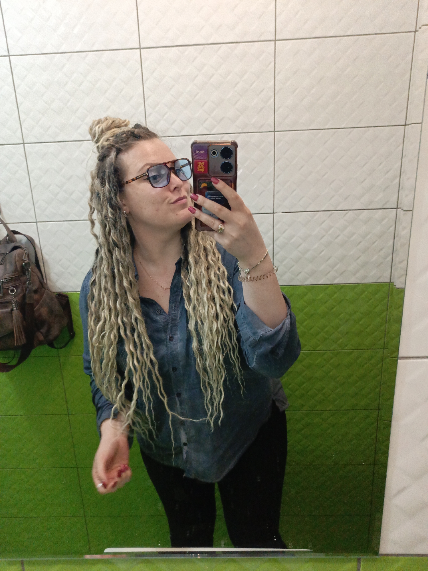 A female is taking a selfie in a bathroom, likely in Verkh-Sosnova, Russia. She has blonde, dreadlock-style hair with some of it pulled into a bun on top of her head. She wears glasses, a blue button-down shirt, and black pants. She has a ring on her finger and a bracelet on her wrist. She is holding a smartphone with a phone case featuring writing and possibly other designs. The bathroom features white textured tiles on the upper portion of the walls and green textured tiles on the lower portion. A brown backpack is hanging on the wall to the left. The image is a reflection in a mirror.