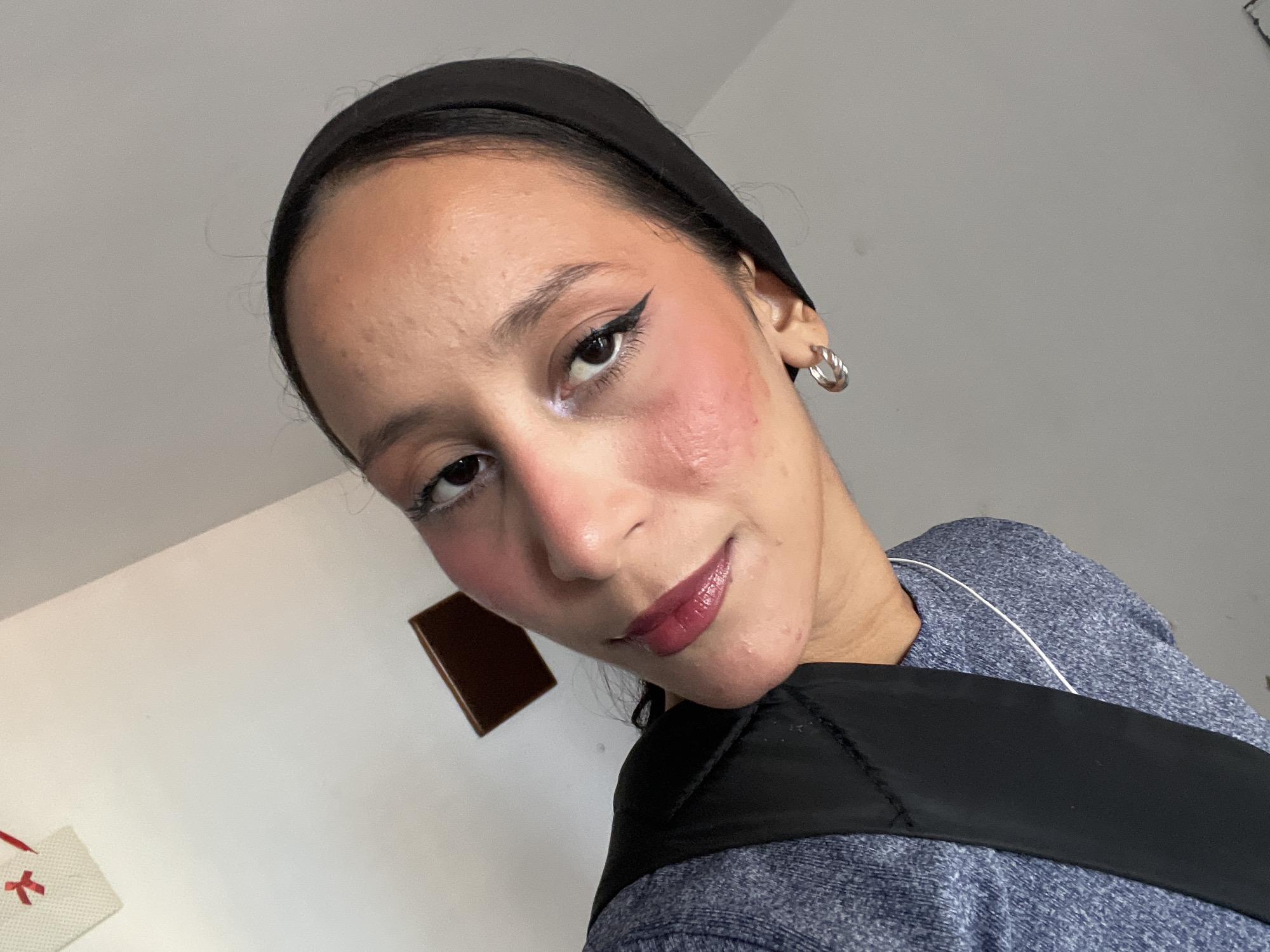 A woman wearing a black hijab and a grey sweater is leaning into the frame, capturing a selfie. Her face is turned slightly to the left, with her right eye visible. She has a bold winged eyeliner and rosy blush applied to her cheeks. Her lips are painted with a deep berry lipstick. She is wearing a silver hoop earring on her visible ear. A black strap is visible across her chest, part of what appears to be a bag or backpack. In the background, a plain light-colored wall is visible, with a small rectangular dark brown object mounted on it. In the upper left corner, a white square with red cross shapes is partially visible. The image does not contain information that can be used to determine its location in Porlamar, Venezuela.
