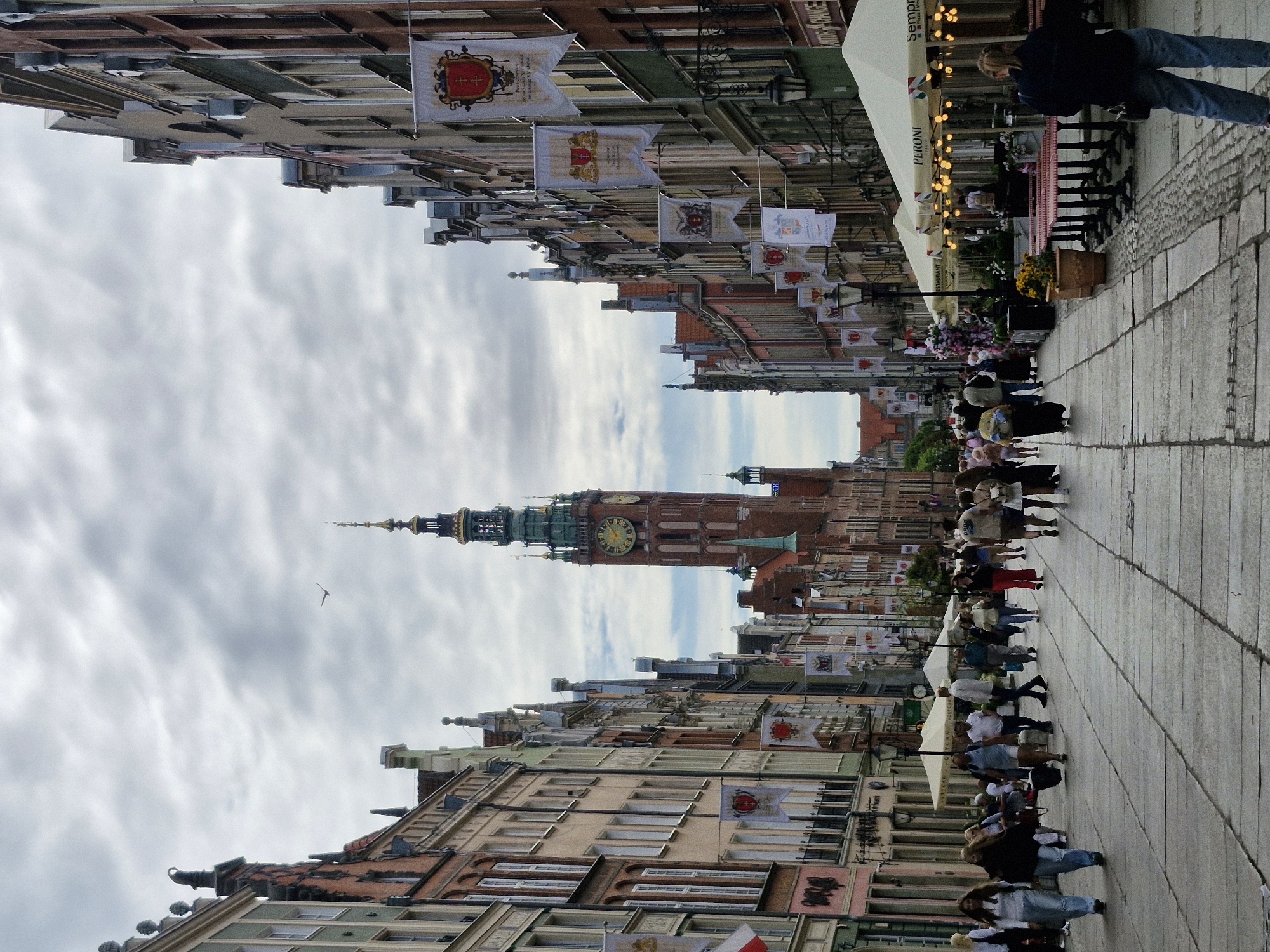 This is a vertical, eye-level, full-shot photograph taken from the street looking down the length of Długa Street in Gdańsk, Poland, towards the Main Town Hall. The street is paved with large, flat, grey stones, with a cobblestone border on the right. The sky is overcast with heavy grey clouds.
On the left and right sides of the street are rows of tall, narrow buildings with distinct architectural styles, featuring pitched roofs, ornate facades, and numerous windows. Many of these buildings are painted in pastel colors, including pink, yellow, and green. Several white banners with coats of arms are hung from the buildings on both sides of the street.
In the distance, the Main Town Hall is prominently visible, a large brick structure with a tall, green spire and a prominent clock face showing it is 3:25. The street is populated with numerous pedestrians walking in both directions. People are dressed in casual clothing, and some are walking in groups. On the right side of the street, several outdoor seating areas for restaurants are set up, with white umbrellas and tables and chairs. Strings of lights are visible under the umbrellas, and there are potted plants and flower arrangements.
On the far left, a woman in a white shirt and blue jeans is walking towards the viewer. To her right, another woman in a black jacket and blue jeans is walking in the same direction. Further down the street on the left, more people are seen walking and milling about. On the right side of the street, a woman in a dark jacket and blue jeans is walking away from the viewer, her back to the camera. Restaurant tables and umbrellas are also visible on this side. In the sky, a bird is flying.