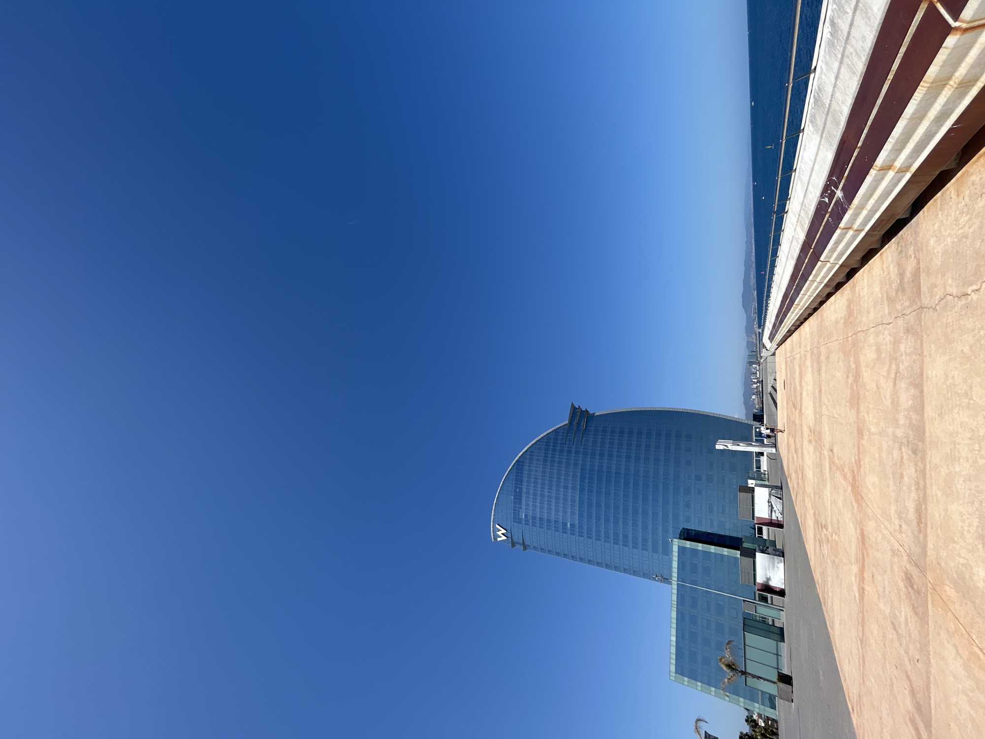 This is a vertical, eye-level, outdoor photograph of the W Hotel in Barcelona, Spain. The hotel, a modern, sail-shaped skyscraper made of blue glass, is the primary subject, dominating the left side of the frame. A large white "W" logo is visible at the apex of the building. In front of the hotel are several smaller, rectangular glass structures, and a single palm tree.
The foreground is a wide pedestrian walkway made of light brown paving stones with a textured, cracked surface. To the right of the walkway, a long, white, concrete structure with dark red stripes runs parallel to the coastline. This structure appears to be a pier or elevated walkway. A metal railing runs along its edge.
In the middle ground, a person in a white shirt and dark shorts walks away from the camera on the paved walkway. Further in the distance, along the coastline, there are several buildings and the silhouette of hills.
The background is a vast expanse of clear, bright blue sky with no clouds. The Mediterranean Sea is visible on the right side of the image, stretching to the horizon. There are a few distant boats on the water. The overall lighting suggests it is a sunny day.