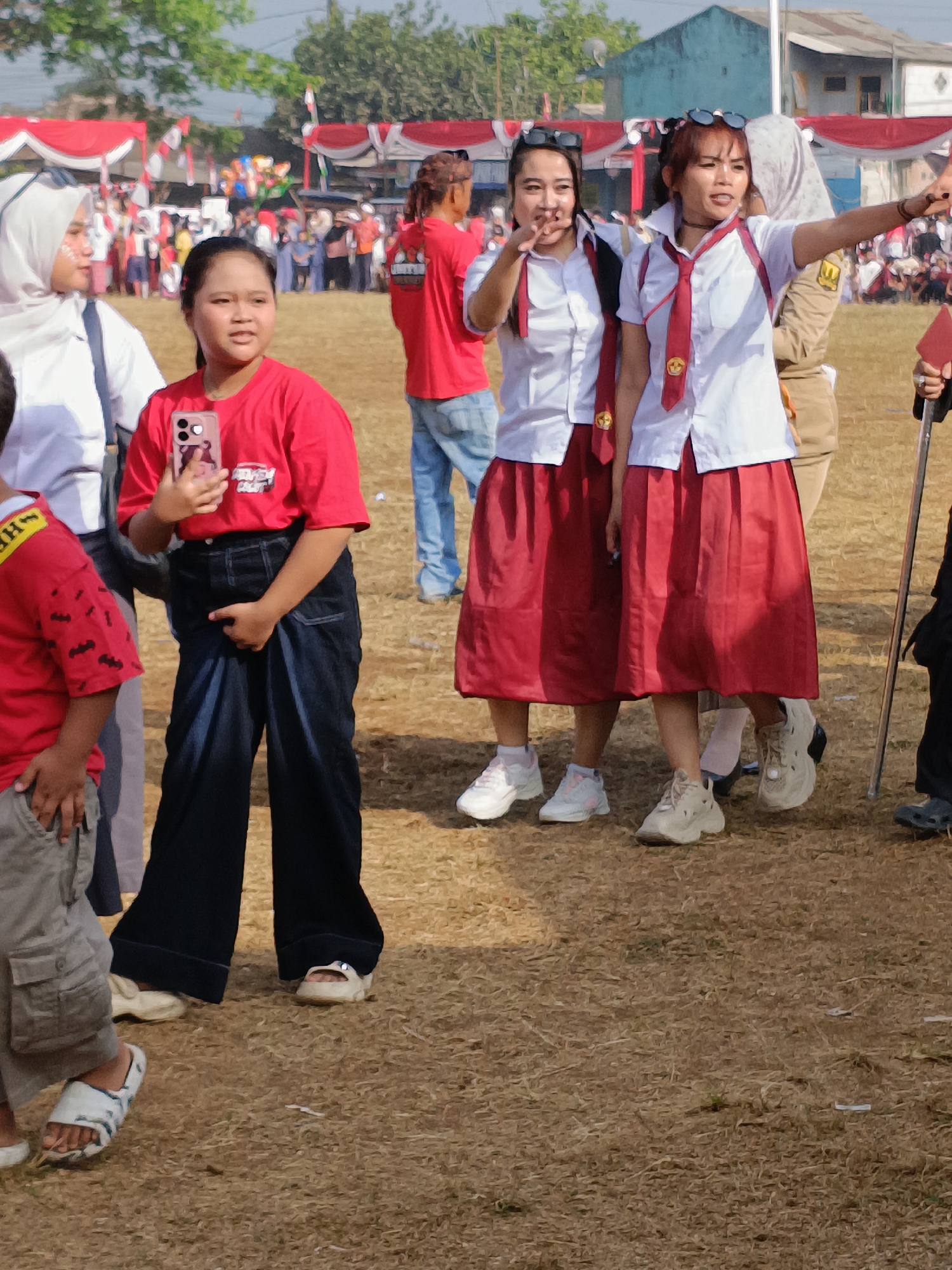 In Cibodas, Indonesia, a group of individuals, predominantly young females, are gathered outdoors on a dry, grassy field under a clear sky. In the foreground, a young girl wearing a red t-shirt and dark, wide-legged pants holds a pink smartphone. To her right, two young women in school uniforms, white shirts and red skirts, stand together. The woman on the right, with sunglasses perched on her head, is gesturing with her right arm outstretched. Behind them, other people are visible, some in red shirts and jeans, and others in what appear to be uniforms. A tent structure decorated with red and white fabric is present in the background, suggesting an event or festival. The overall scene depicts a casual outdoor gathering with a festive atmosphere.