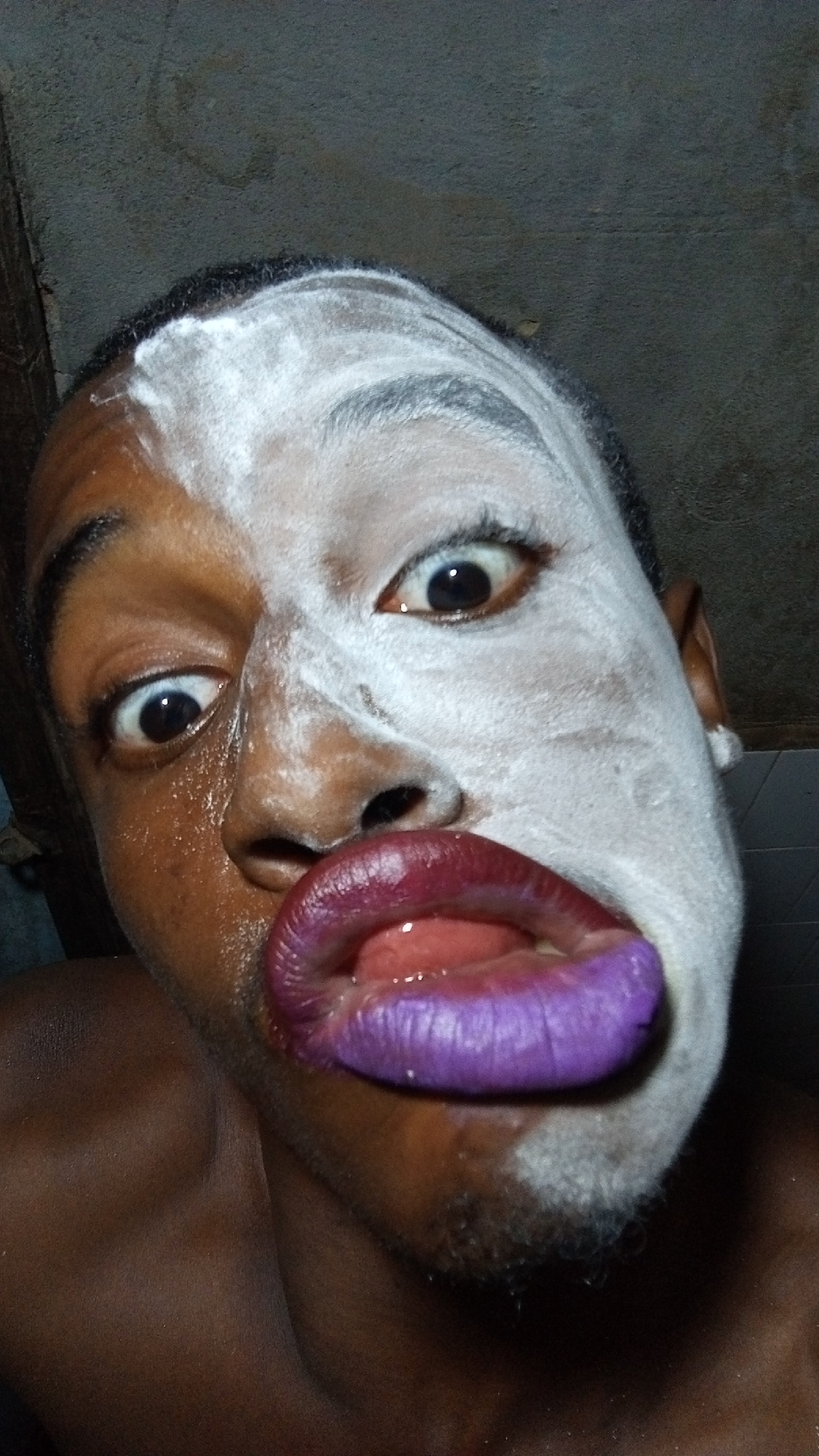 A close-up, low-angle shot captures a person with their face partially covered in a white substance. The right side of their face is coated in this white material, while the left side reveals dark skin. Their lips are prominently featured and are colored a vivid purple. The person's eyes are wide and appear somewhat startled. The background is indistinct, possibly an interior space with neutral-toned walls.
