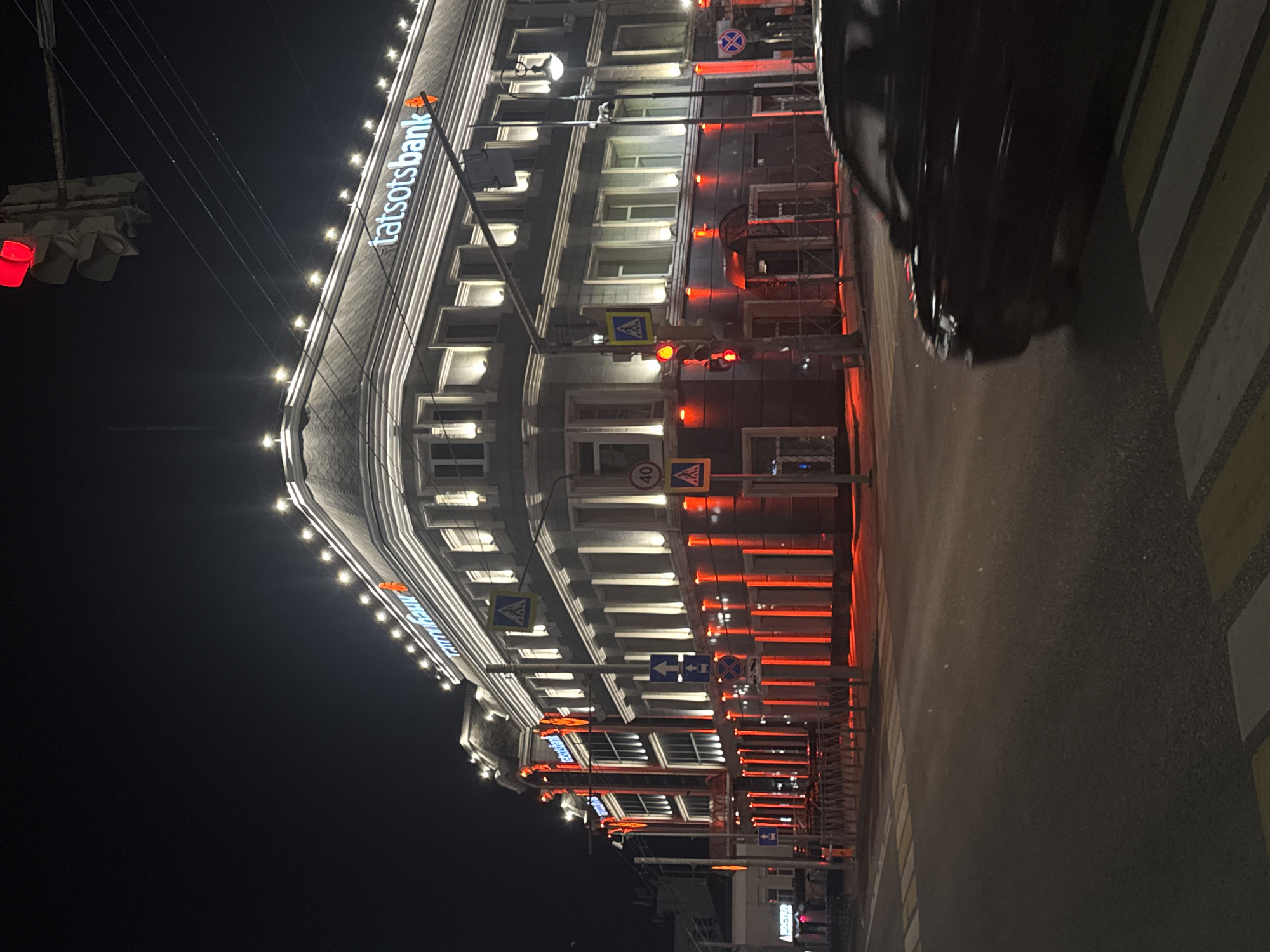 This is a nighttime, medium-angle, eye-level exterior shot of the Tattsotsbank building in Kazan, Russia. The building, an older, multi-story structure with classical architectural elements, is illuminated by bright red lights along its lower facade and by numerous small, warm white lights along its roofline and cornices. The sign for "tatstsotsbank" is prominently displayed in large, illuminated white letters on the building's upper facade. A traffic light with a red signal is visible in the upper left of the frame. Several street signs, including pedestrian crossing signs and directional arrows, are present. In the foreground, a dark-colored car is blurred as it drives across a pedestrian crossing marked with yellow and white stripes. The street and sidewalk are visible, with the latter featuring a metal railing. The sky is dark, indicating nighttime.