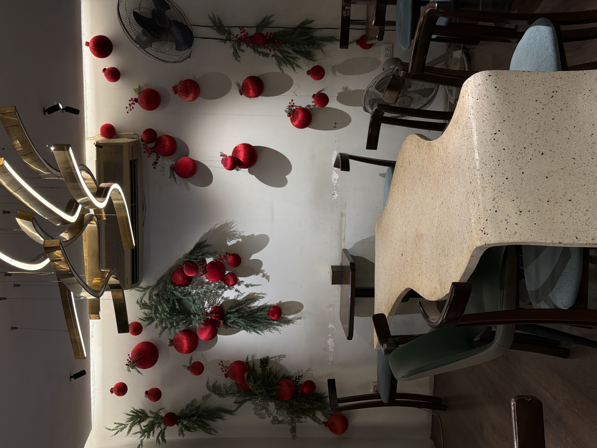 This interior shot depicts a festive setting, likely within a cafe or restaurant in Hà Nội, Vietnam. The scene is dominated by a wall extensively decorated with holiday-themed elements: numerous red, velvety spherical ornaments, some adorned with small red berries, are interspersed with green artificial pine or fir branches. Shadows cast by these decorations suggest directional lighting.
On the left side of this decorated wall, a modern, sculptural light fixture with golden, wavy elements and integrated LED strips provides illumination. Below it, a white air conditioning unit is mounted, and further left, two small black spotlights are visible. Another wall-mounted fan is positioned high on the wall in the upper right corner, and a second fan is partially visible towards the lower right.
To the right of the decorated wall, a dining area is set up. A prominent table with a light-colored, speckled (terrazzo-like) surface extends across the right half of the frame. Several chairs with dark wooden frames and light blue or green upholstered seats are arranged around the table. The lighting in the room is artificial, creating a cozy but slightly dim ambiance, suggesting evening or nighttime. No people are present, indicating a quiet moment in the establishment. No visible text is discernible.