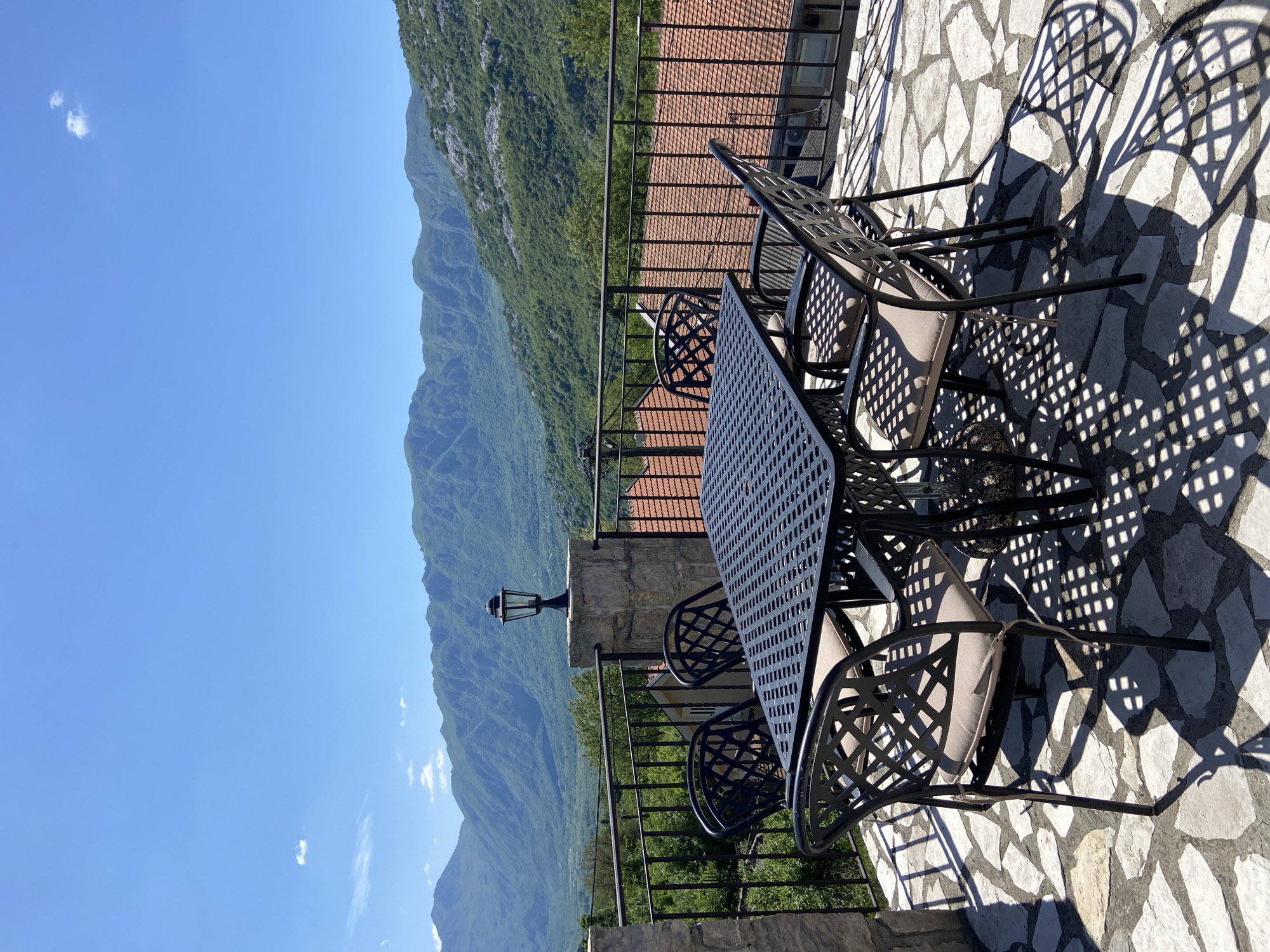 The image captures a sunny outdoor patio or balcony in Sotonići, Montenegro, offering a stunning panoramic view of a mountainous landscape. In the foreground, a dark metal patio table with a perforated surface is surrounded by four matching chairs, each featuring an intricate lattice design and light-colored cushions. Strong, sharp shadows from the furniture are cast across the irregular, light-toned stone paving of the patio floor, indicating bright midday sun.
A black metal railing runs across the midground, separating the patio from the expansive vista. A small, classic lantern-style lamp post stands on a stone pillar near the railing. Beyond the railing, a lush, verdant valley stretches out, giving way to majestic, rolling mountains covered in dense greenery. The sky above is a clear, vibrant blue with only a few small, wispy white clouds. Below the patio level, parts of a building with a distinct red-tiled roof are visible, nestled into the landscape.
No people are present, suggesting a peaceful and serene setting, ideal for relaxation or dining al fresco. The weather is clear and sunny, enhancing the vibrant colors of the mountains and sky. There is no visible text in the image.