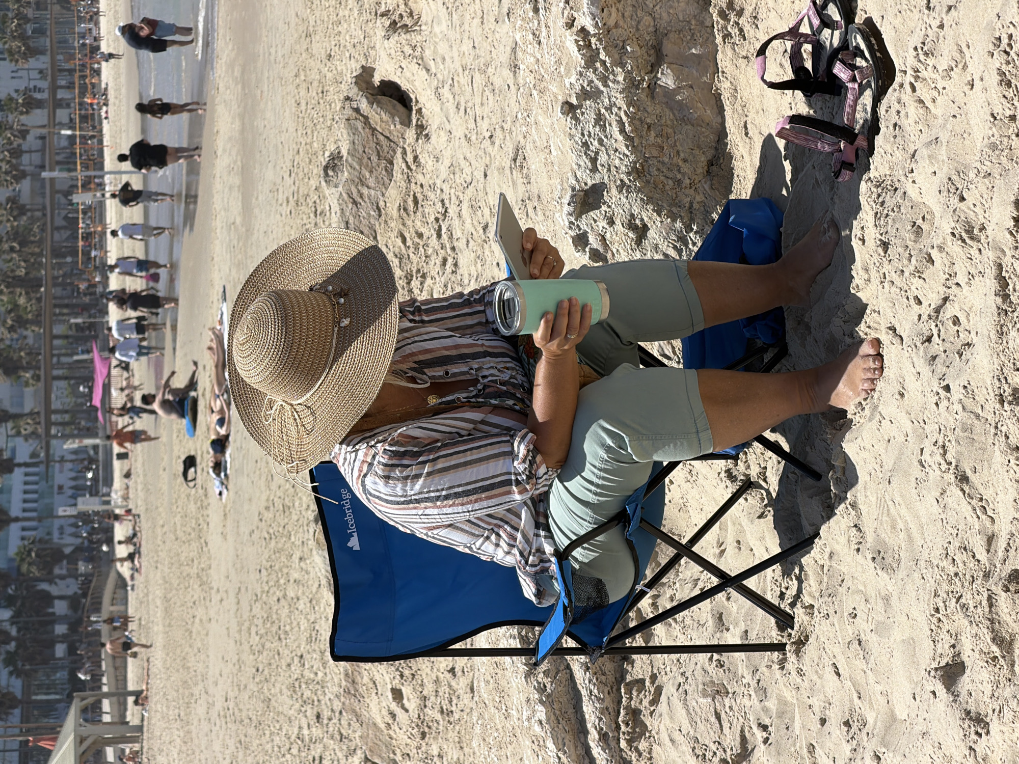 A woman sits in a blue folding chair on a sandy beach, facing right. She wears a straw hat, a striped shirt, and green shorts. In her hands, she holds a phone and a light green water bottle. To her right, on the sand, are pink and black straps. Behind her, the beach is visible, populated with people, buildings, and palm trees. The beach and buildings suggest the location is Tel-Aviv, Israel. The background scene depicts a typical beach setting with people sunbathing, walking, and swimming.