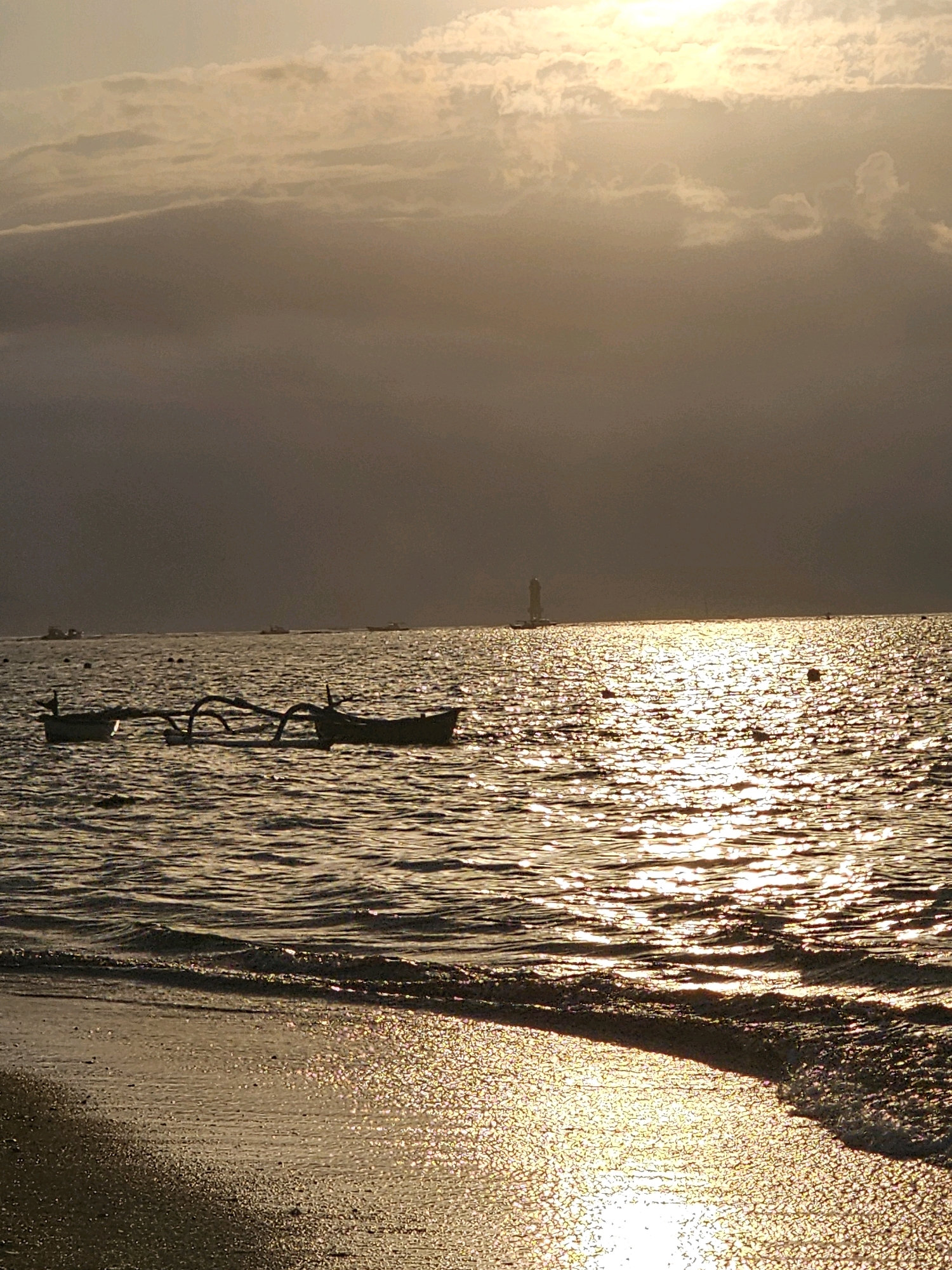 The image depicts a serene seascape at sunset, likely on a beach in Denpasar, Indonesia, given the presence of traditional outrigger boats. The sun is setting, casting a warm, golden glow across the water, creating shimmering reflections. In the foreground, gentle waves lap onto a sandy shore. Two outrigger boats are visible in the mid-ground, bobbing on the water. In the distance, a faint silhouette of a lighthouse can be seen against the horizon, along with other small boats. The sky is partly cloudy, with the sun partially obscured by a band of clouds, contributing to the atmospheric lighting. The overall mood is tranquil and picturesque.