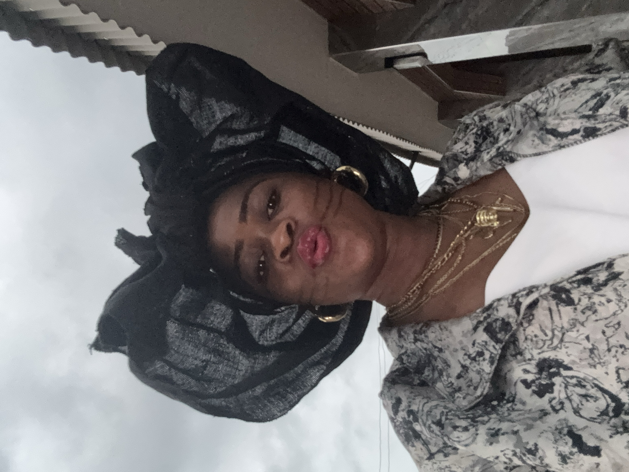 A low-angle, medium shot captures a Black woman from the shoulders up, standing outdoors in the city of Ajara, Nigeria. The woman is wearing a black fascinator hat adorned with large, textured fabric bows. She also wears multiple gold necklaces, one with a large, gold pendant shaped like an insect, and large gold hoop earrings. Her lips are pursed, as if for a kiss, and she is wearing makeup. Her patterned jacket, in shades of black, white, and grey, is open over a white top. Behind her, the sky is overcast, and part of a building with a textured, grey roof and a light-colored wall can be seen to the right of the frame. The overall scene suggests a formal or festive occasion.
