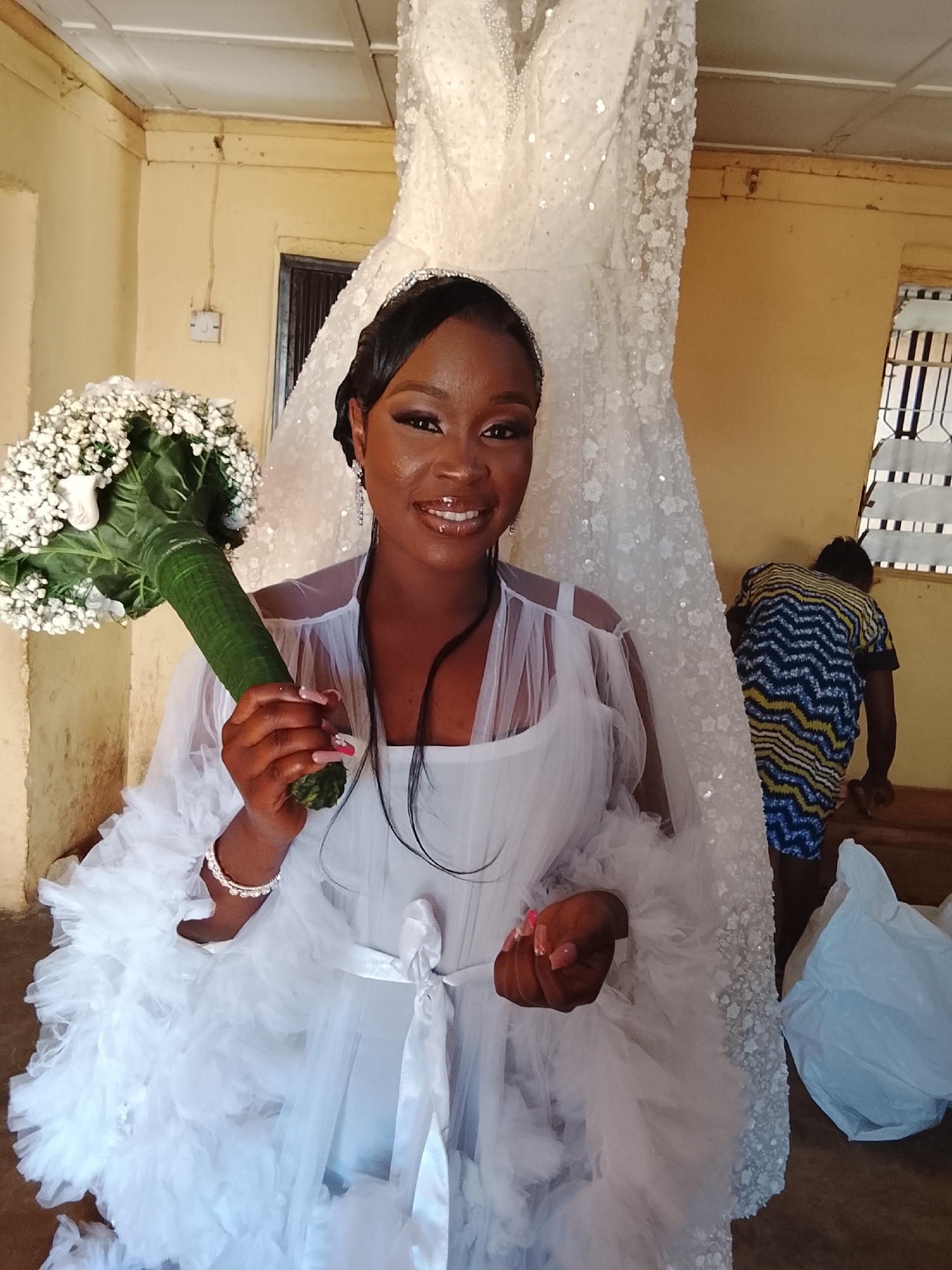 The image shows a dark-skinned woman, presumably a bride, holding a bouquet of white flowers. She wears a white robe over a white garment, a headband, and earrings. A wedding gown is hung behind her. In the background, a person in a patterned blue, yellow, and white shirt is visible, along with a white plastic bag. The location appears to be indoors with yellowish walls. The location in the city is unknown.