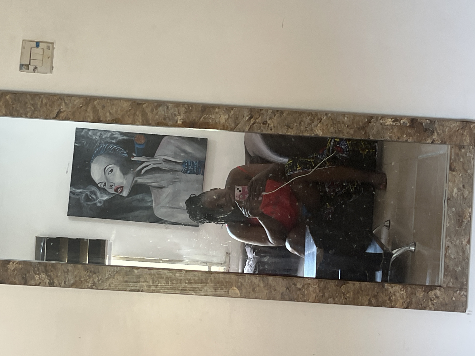 A full-length mirror in a room reflects a woman seated on a sofa, holding a smartphone. The woman has braided hair and is wearing a red top and a patterned skirt. A cable is connected to her phone. Behind her, a painting of a woman smoking a cigar is visible on the wall. The room appears to be in an apartment in Abuja, Nigeria, as indicated by the electrical outlet on the wall, which is consistent with outlets commonly found in Nigeria.
