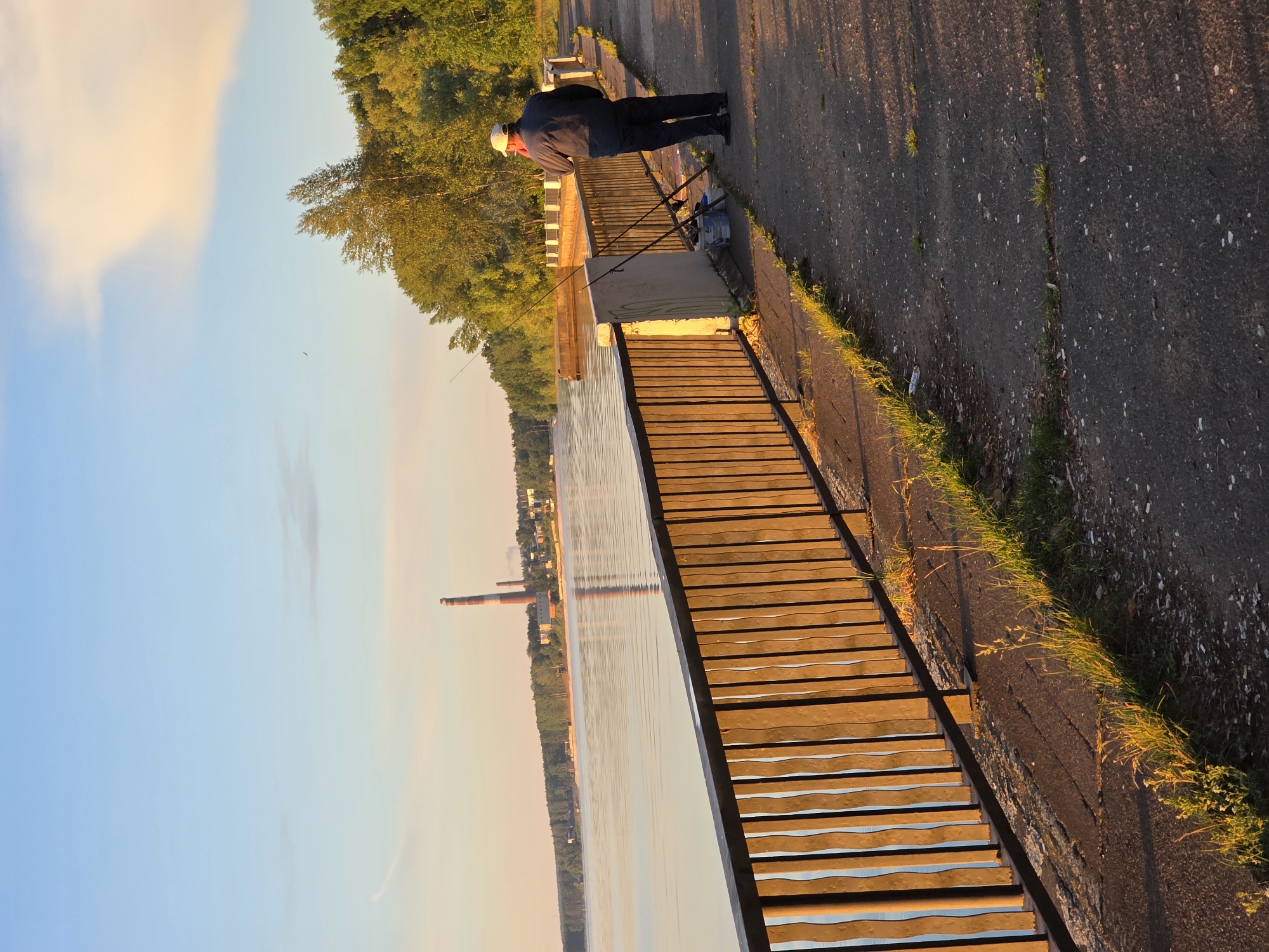 An elderly man stands on a paved embankment, facing a body of water, at the city of Chaikovsky, Russia. He is wearing a dark jacket, dark trousers, and a light-colored cap. Two fishing rods are propped up beside him, along with a bucket. A wooden fence runs along the embankment, separating it from the water. In the distance, across the water, is a shoreline lined with trees and buildings, including a tall, striped industrial chimney. The sky is a soft blue with streaks of pale orange and yellow clouds, indicating either dawn or dusk. The light suggests the golden hour.
