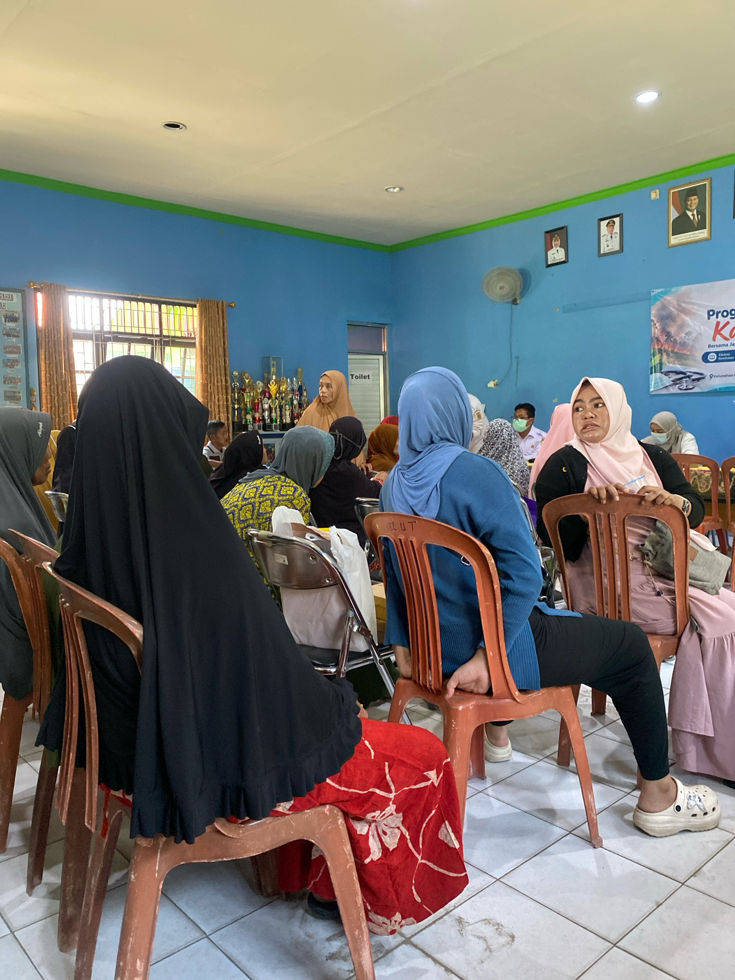 In Banjarbaru, Indonesia, a group of women are seated in a room, likely a community hall or classroom, attending an event. The room has blue walls and a white ceiling with fluorescent lighting. The women are dressed in modest Islamic attire, with many wearing hijabs. Some women are looking towards the front of the room, where a speaker might be situated, while others are engaged in conversation. In the background, there are framed pictures on the wall, a collection of trophies, and a banner with Indonesian text. The floor is tiled. One woman in the foreground is wearing a black abaya and a red patterned skirt, while another in a blue hijab is seated and looking to her right. The overall atmosphere appears to be one of a gathering or presentation.