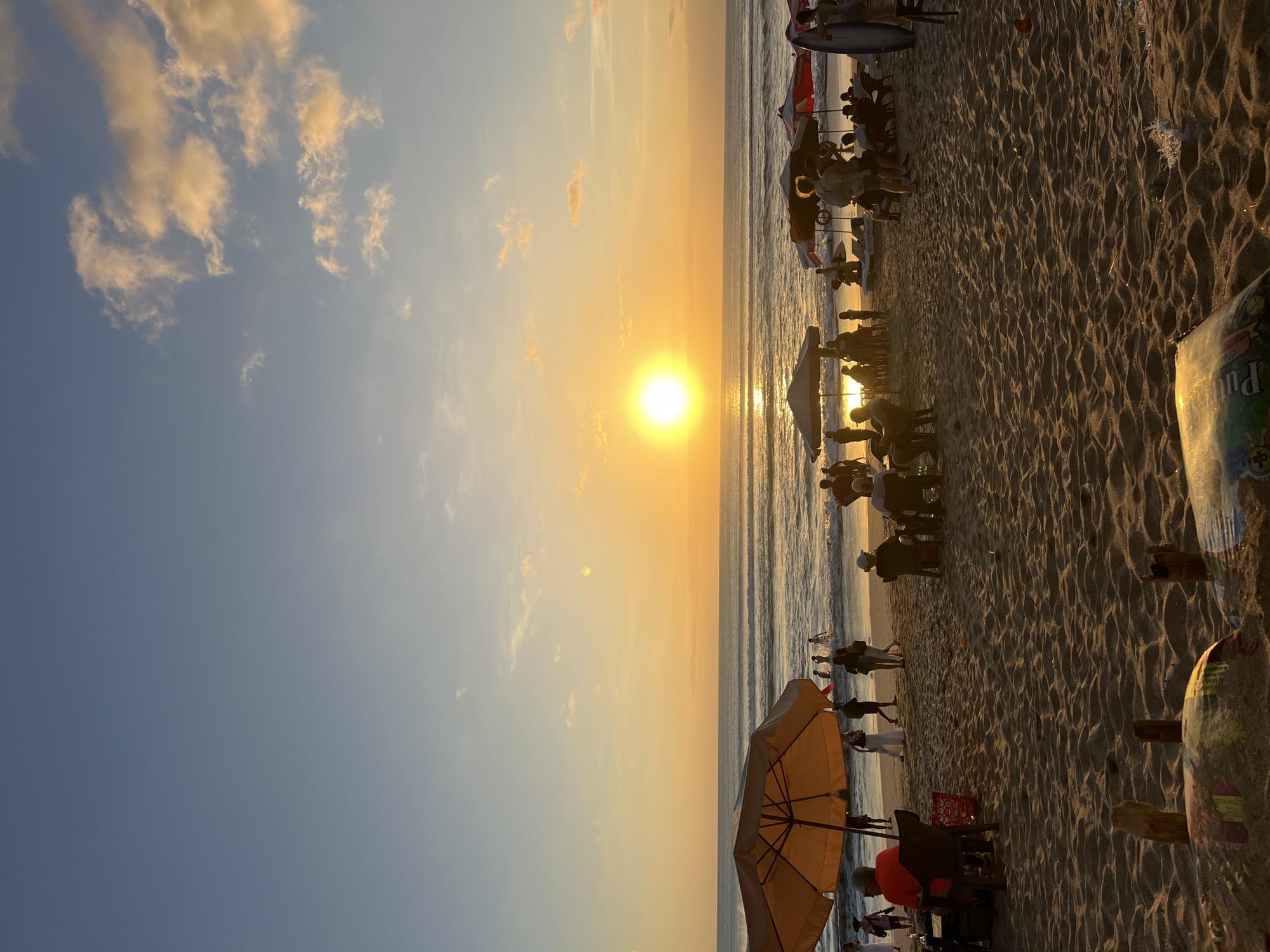 This image captures a vibrant sunset scene on Kuta Beach, Bali, Indonesia. The sun, a brilliant orb of golden light, is setting over the ocean, casting a warm glow across the sky and water. Gentle waves roll onto the sandy shore, where numerous people are gathered.
The beach is dotted with beach umbrellas, some providing shade for beachgoers, while others are set up near small tables. People are seen sitting, standing, and walking along the shore. Some are enjoying the view, while others appear to be engaged in conversation or simply relaxing. A few individuals are wading in the shallow water, enjoying the refreshing ocean breeze.
In the foreground, the sand is textured with footprints and the remnants of beach activities. Some scattered debris, including plastic bags, are visible on the sand. The overall atmosphere is one of leisure and tranquility, with the natural beauty of the sunset providing a stunning backdrop to the human presence on Kuta Beach.