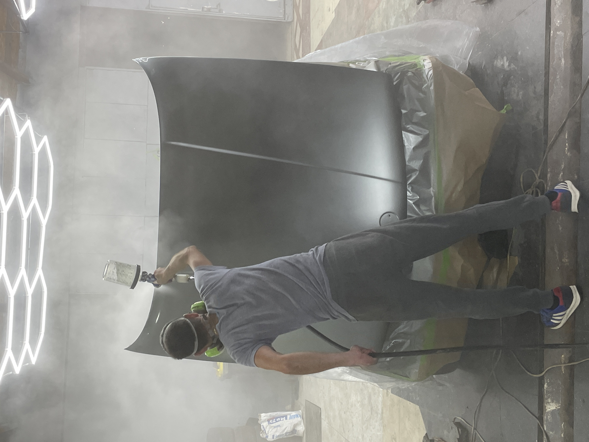 A person is spray painting a car hood in a dimly lit workshop. The hood is dark gray and is propped up on supports. The person is wearing a grey t-shirt, dark pants, and green ear protection. They are holding a spray gun and are in the process of applying paint to the hood. There is a lot of overspray in the air, indicating the painting process is underway. In the background, there are hexagonal light fixtures on the wall, casting a white glow. There is also a large roll of plastic sheeting and some brown paper covering parts of the surrounding area, likely to protect them from paint. A white bag with blue text is visible on the floor, it reads "KNAUF MP75". The scene appears to be an indoor auto body shop or a similar workspace.