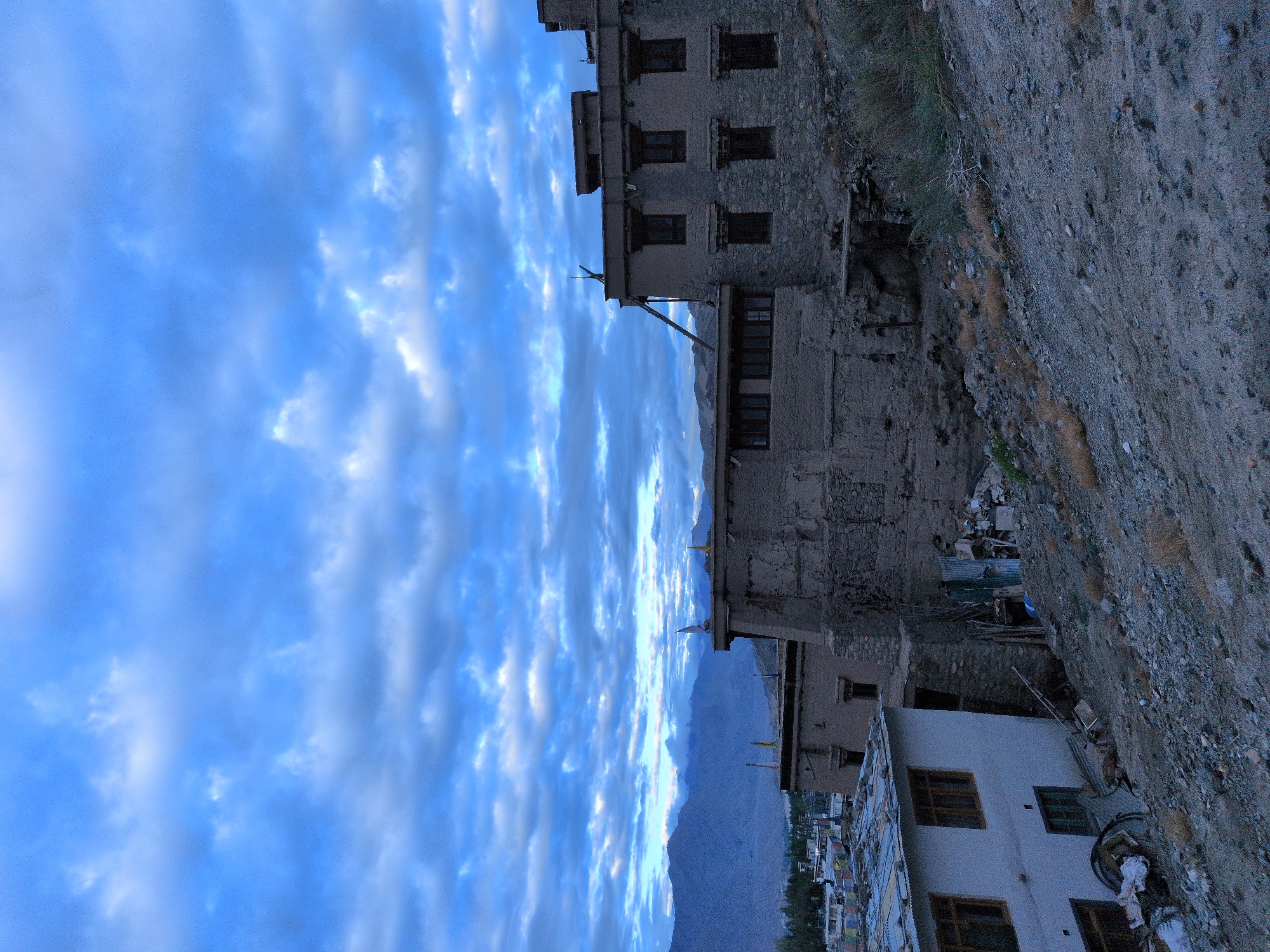 In the city of Leh, India, at dusk, a multi-story ancient Tibetan-style building constructed of stone and mud sits on a hill, with other smaller structures and prayer flags visible in the distance. The sky is cloudy and blue, with mountains in the background. The foreground consists of a rocky, sloped terrain.