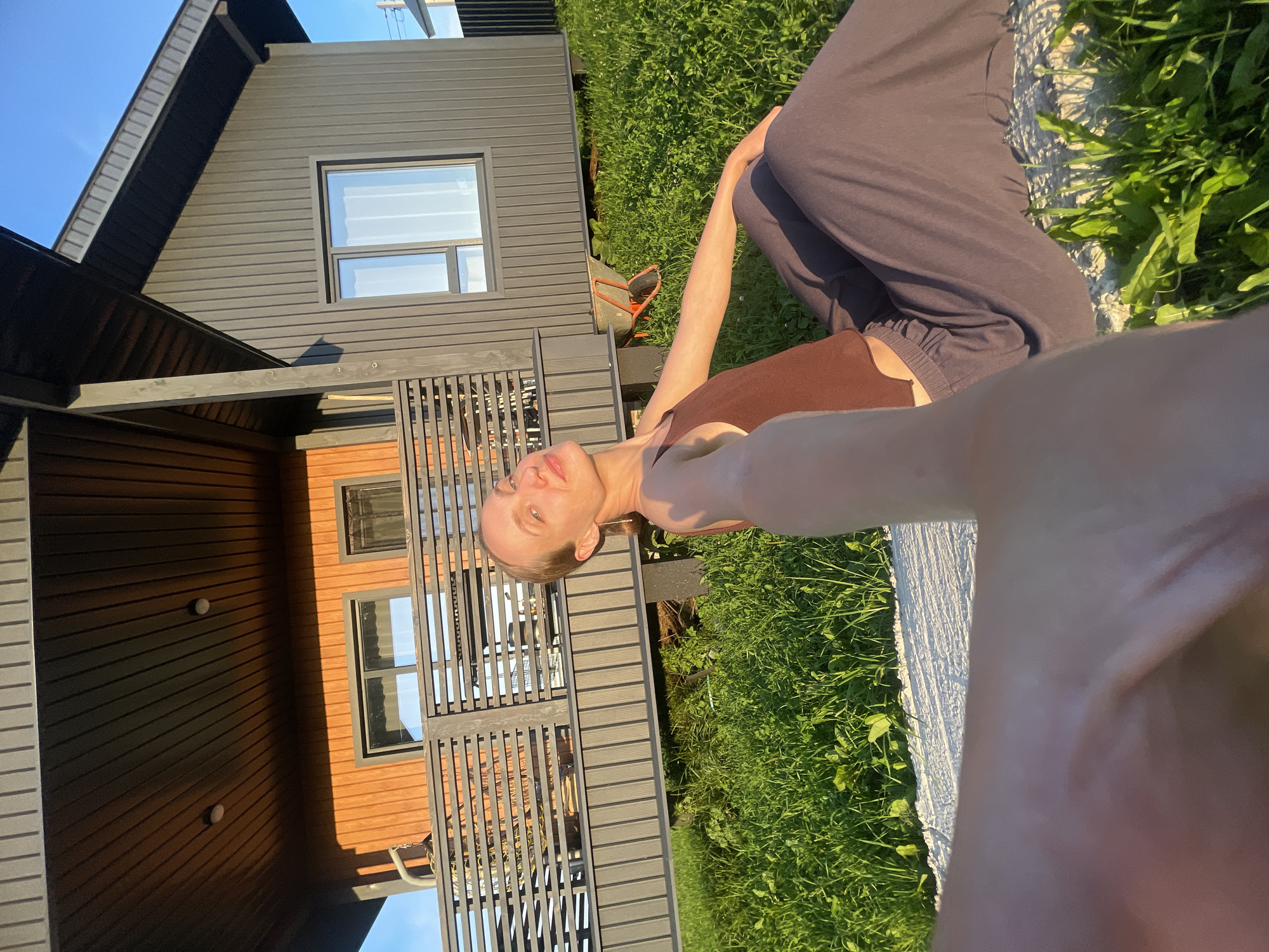 A blonde woman with fair skin, wearing a brown tank top and dark grey pants, is sitting on the grass in front of a modern, dark grey and orange wooden house. The house has a covered porch with a wooden railing and two windows visible. The sun is setting, casting long shadows and illuminating the scene with warm golden light. A wheelbarrow is visible in the background on the grass. The woman is looking towards the camera, with her arm extended as if taking a selfie. The environment appears to be rural, with a clear blue sky. The location is identified as Podshivalovo, Russia.