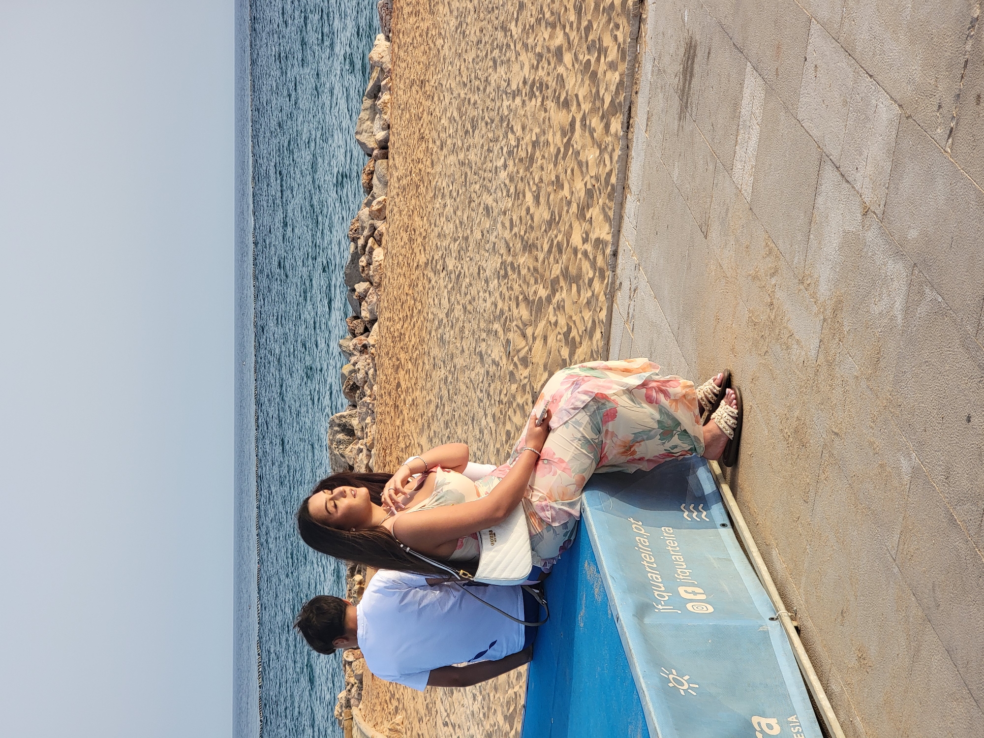 Two individuals are seated on a blue structure near the beach in Quarteira, Portugal. A woman, facing to the right and with her eyes closed, wears a floral-patterned dress and a white purse. She has jewelry on her wrists and a ring on her right hand. To her left and further back, a person in a white t-shirt is seated with their back to the camera. The background features a sandy beach with rocks, the ocean, and a clear sky. Text on the blue structure includes "jf-quarteira.pt" and "jfquarteira".