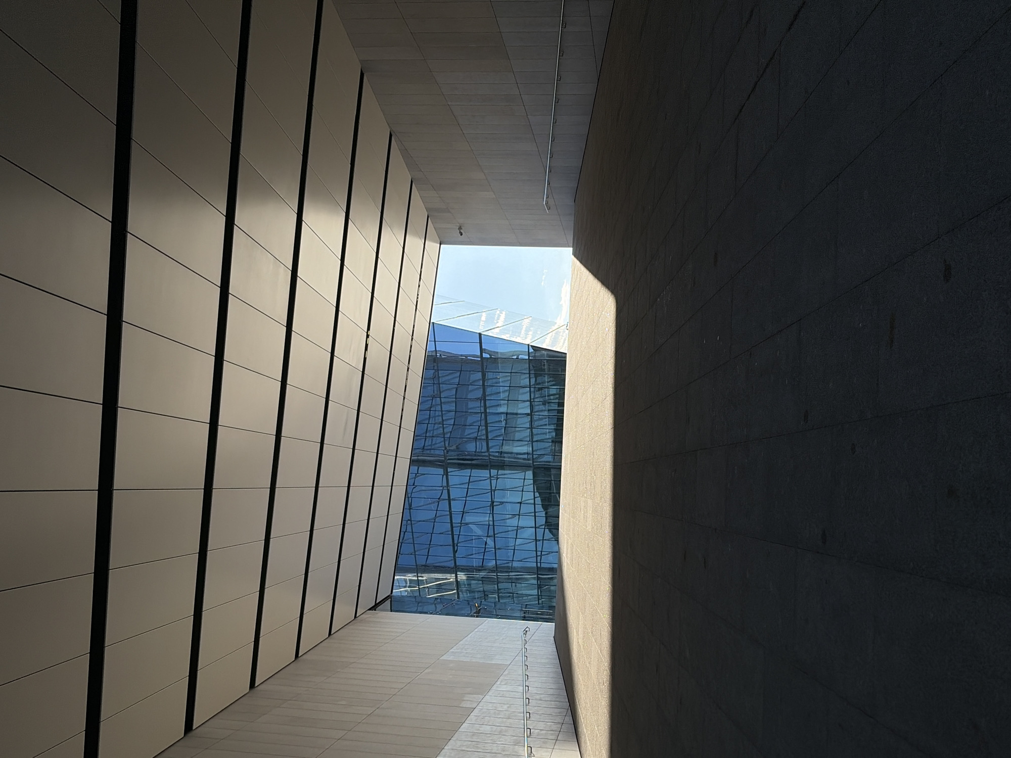 The image depicts a modern architectural perspective of a walkway leading towards a glass-fronted building. The walkway has a tiled floor and is covered by a ceiling with a grid of light-colored panels and dark linear accents. Sunlight casts a strong shadow across the foreground. To the left and right, the walkway is bordered by light-colored walls. In the distance, the imposing glass facade of a building reflects the blue sky and surrounding architecture, with a prominent angular design. A metal handrail runs along the wall on the right side, and a security camera is visible on the upper right wall. The scene suggests an urban environment with a focus on contemporary design. This is the area in front of the European Central Bank in Frankfurt, Germany.