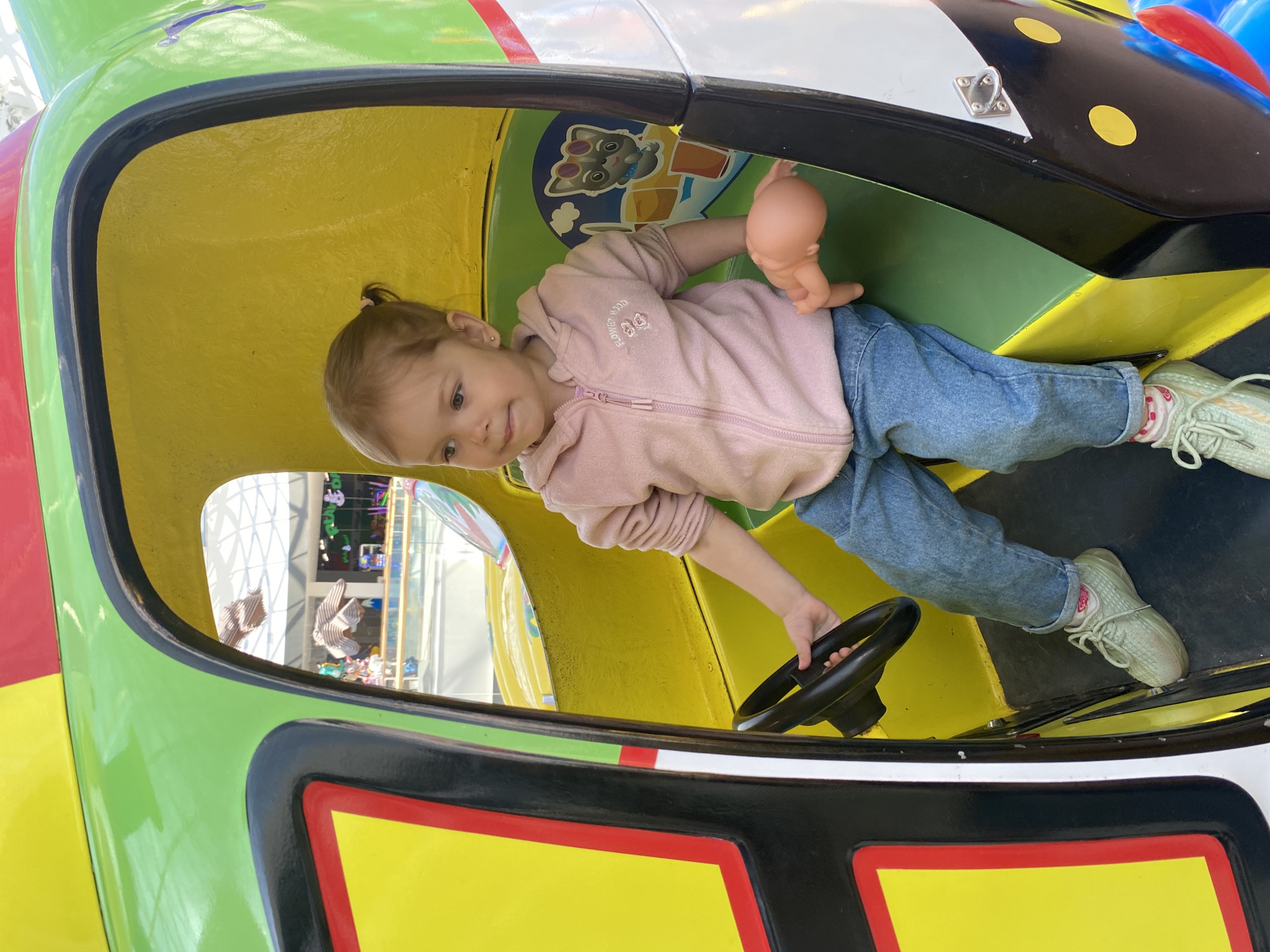 A young girl is seated inside a colorful, cartoon-like vehicle, possibly a children's ride. She is wearing a pink hooded jacket, jeans, and light green sneakers with pink socks. Her hair is pulled back in a ponytail. She is holding a small, nude doll in one hand and gripping a black steering wheel with the other.
The vehicle has a yellow interior and is decorated with cartoon images. The exterior features a combination of green, yellow, red, black, and white paint. A window in the vehicle shows a brightly lit interior space with tall windows and other figures in the background.
Based on the surrounding architecture and distinctive sculptures visible through the window, the location is likely the "iMall Esplanade" shopping center in Perm, Russia.