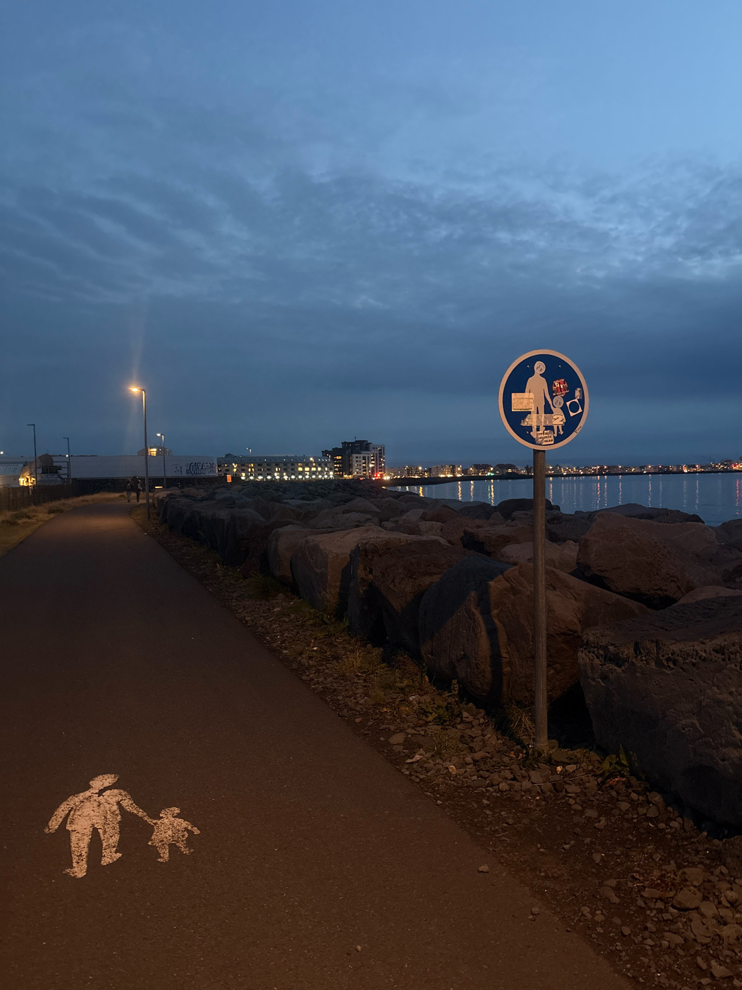 An outdoor, ground-level, eye-level, wide-angle shot captures a path alongside a rocky coastline at dusk in Reykjavik, Iceland. A light-colored, stenciled image of an adult and child walking hand-in-hand is painted on the dark asphalt path. A blue, circular sign with a white border, depicting a person holding bags, a child in a stroller, and a dog, is mounted on a metal pole to the right of the path. The sign is positioned in front of large, dark rocks that border the path. Beyond the rocks is a body of water, reflecting the lights of a city skyline in the distance. The sky above is a deep blue, with faint, streaky clouds. The scene is illuminated by streetlights, casting a faint glow on the path.
