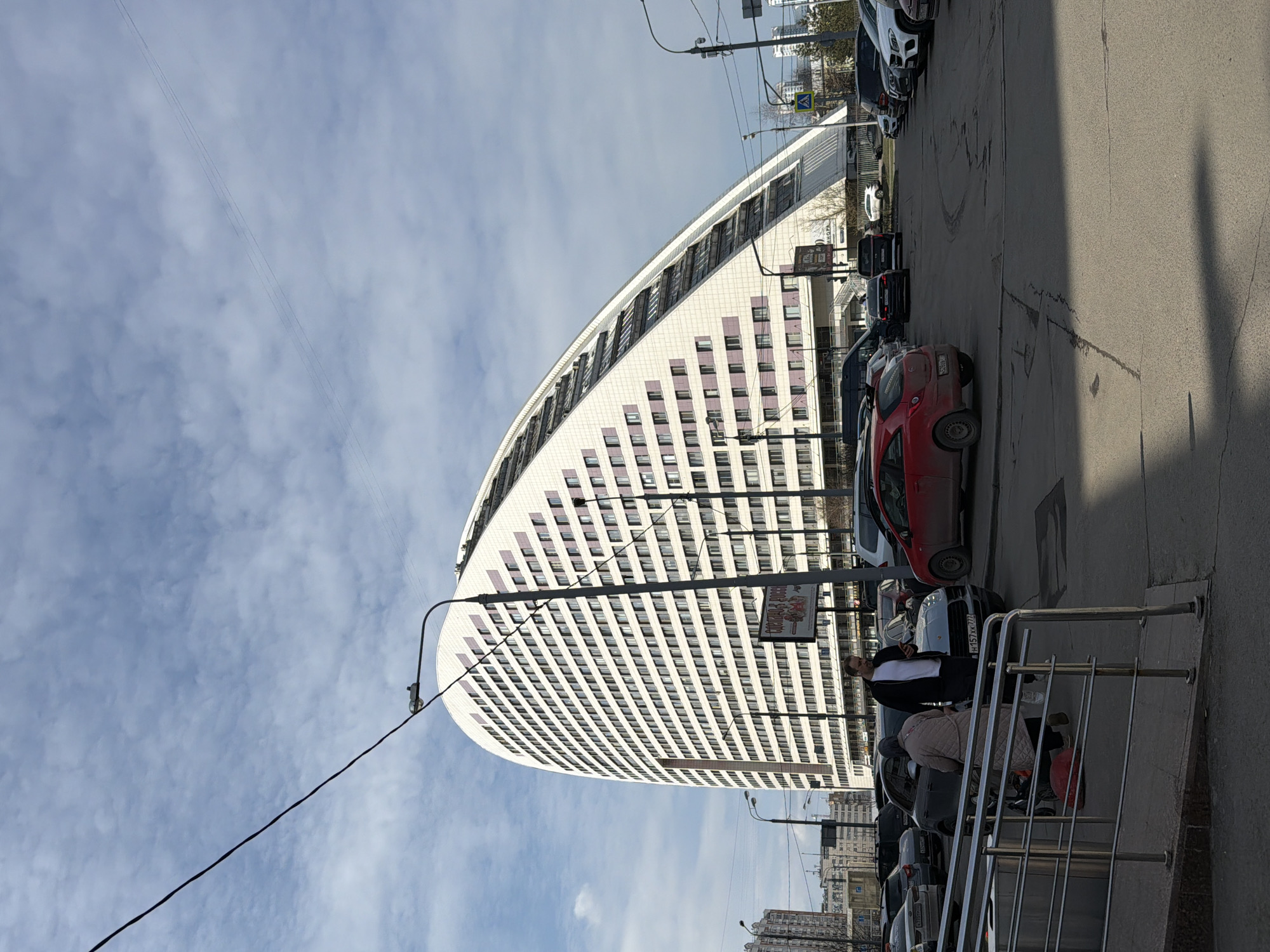 The image depicts a cityscape in Moscow, Russia. A large, modern building with a distinctive curved façade and white-and-grey tiling dominates the frame. The building has many rows of windows and curves to the left.
In the foreground, a street scene is visible, with cars parked along the side. A metal railing runs along the right side of the frame, with people standing and sitting nearby. The people appear to be interacting in a casual setting. Power lines crisscross the sky above. The weather appears to be sunny, with a blue sky and scattered clouds.