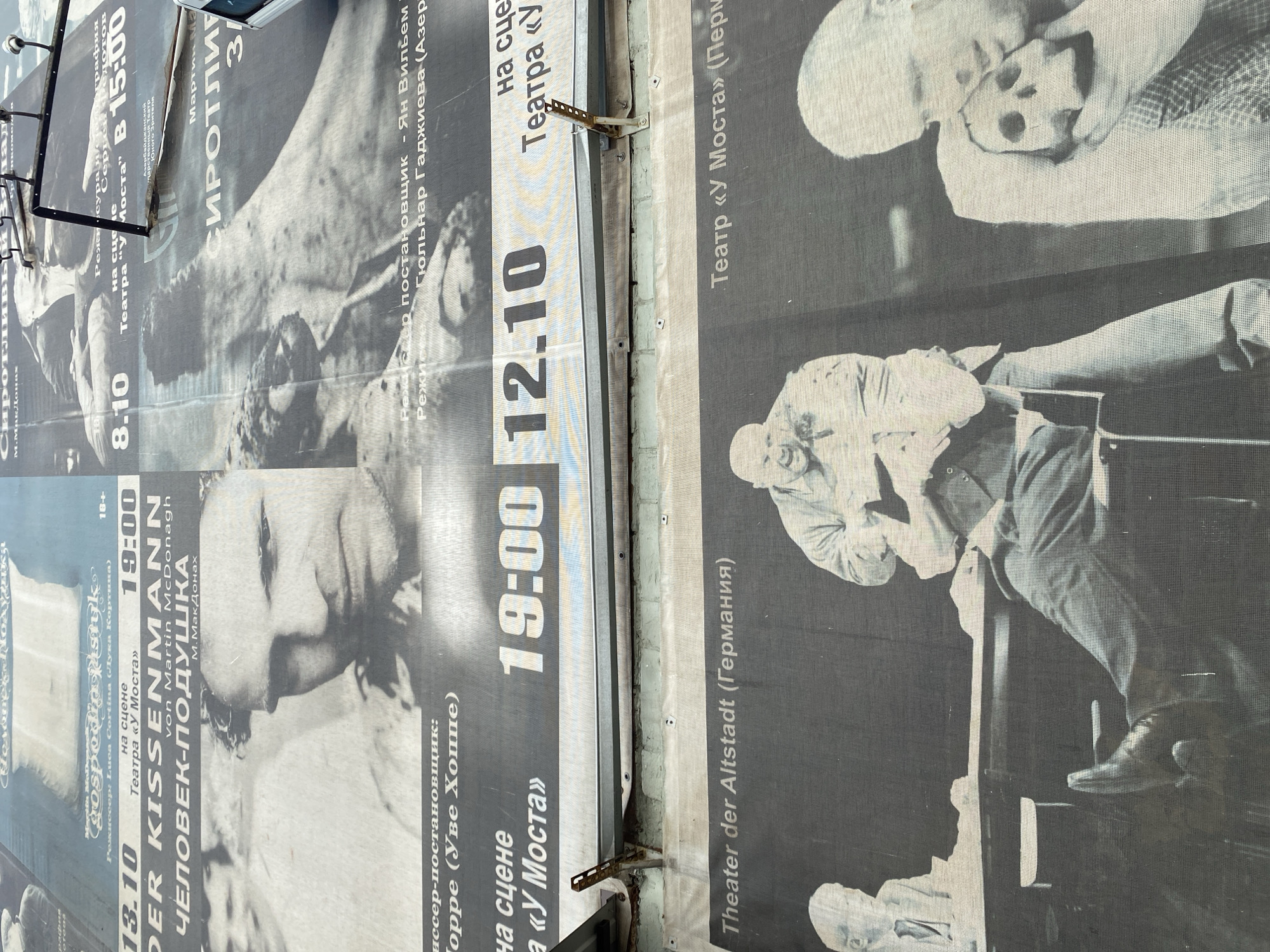 The image displays a large, outdoor billboard advertising a theatrical performance titled "The Pillowman" (Человек-подушка) by Martin McDonagh. The billboard is divided into several sections. The top section features a black and white photograph of two male actors' faces, with one in sharp focus. Text below identifies the play's title in both Russian and English, along with the theater's name, "Театр «У Моста»" (Theater "U Mosta"), which is located in Perm, Russia. Performance dates and times are prominently displayed: "13.10" at "19:00" and "12.10" at "19:00". Further text details the director and other creative team members.
The lower portion of the billboard shows a black and white photograph of actors on a stage, depicting a scene from the play. One actor is seated with his legs crossed, and another is standing, holding a skull. The text "Theater der Altstadt (Германия)" is visible, indicating a collaboration or guest performance from a German theater. The billboard is attached to a brick wall and appears to be weathered, with some damage to the material and mounting hardware visible. The lighting suggests an outdoor, daytime setting.