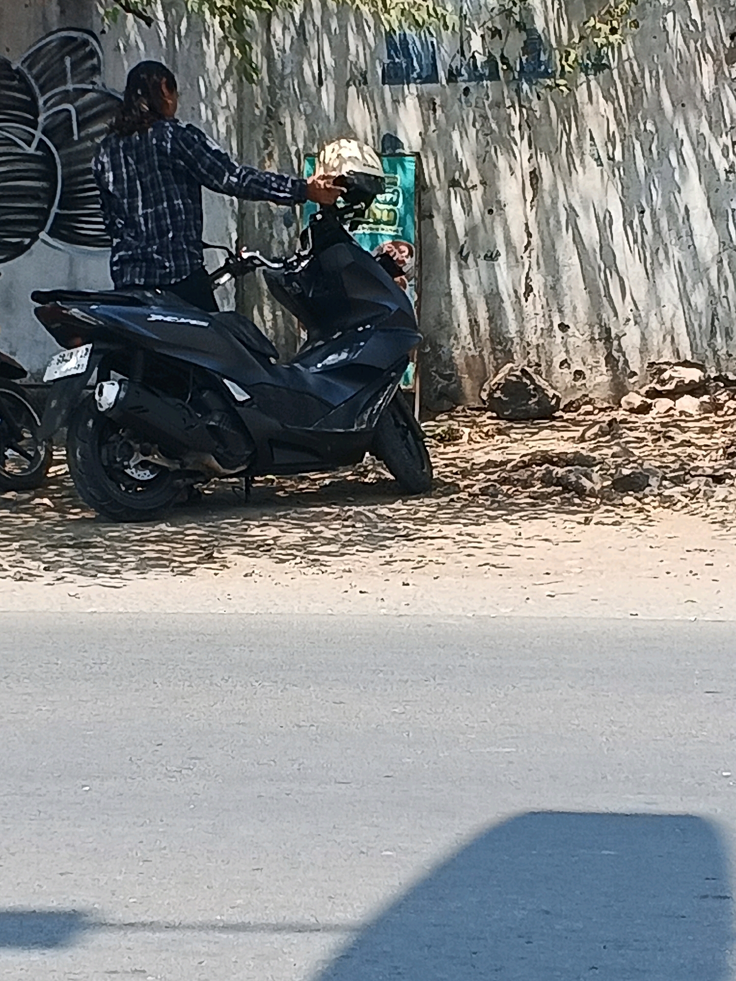 In Taman, Indonesia, a person with long hair, wearing a blue and white plaid shirt, is leaning on a black scooter. The scooter has a license plate that reads "S 2713 YN". The person's right hand is on the scooter's handlebars. A helmet, possibly a white and gold patterned one, is on the back of the scooter. The scene takes place outdoors, with a textured wall in the background that appears to have graffiti on the left side. The ground is covered in dirt and small stones. Sunlight casts shadows on the wall, suggesting it is daytime. The image is taken from a low angle, with a grey paved surface in the foreground, possibly a road.