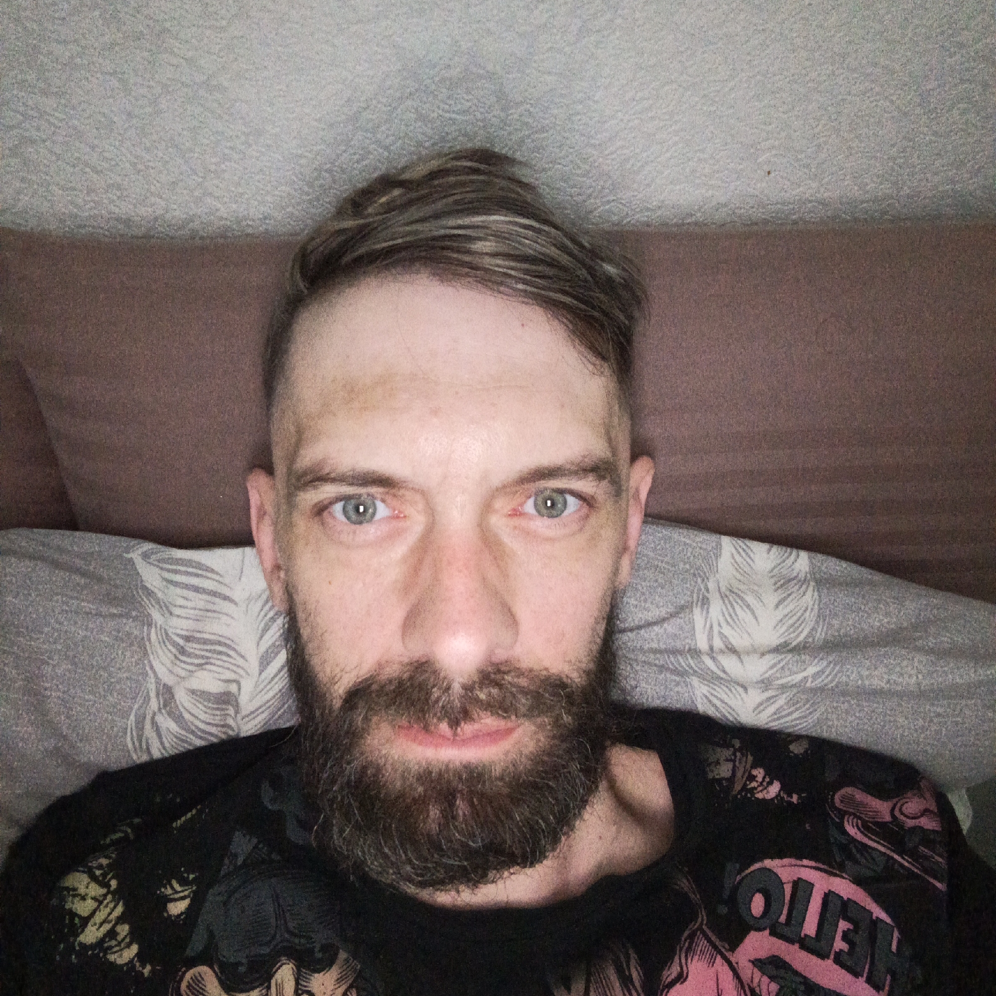 The image depicts a fair-skinned man with a beard lying in bed. He has short, styled hair with lighter streaks. His eyes are light-colored. He's wearing a black t-shirt with a design on it. He appears to be indoors, lying on a patterned pillow with a headboard behind him, and in the background is a textured wall. There is no evidence to suggest the location is Марковский, Russia.