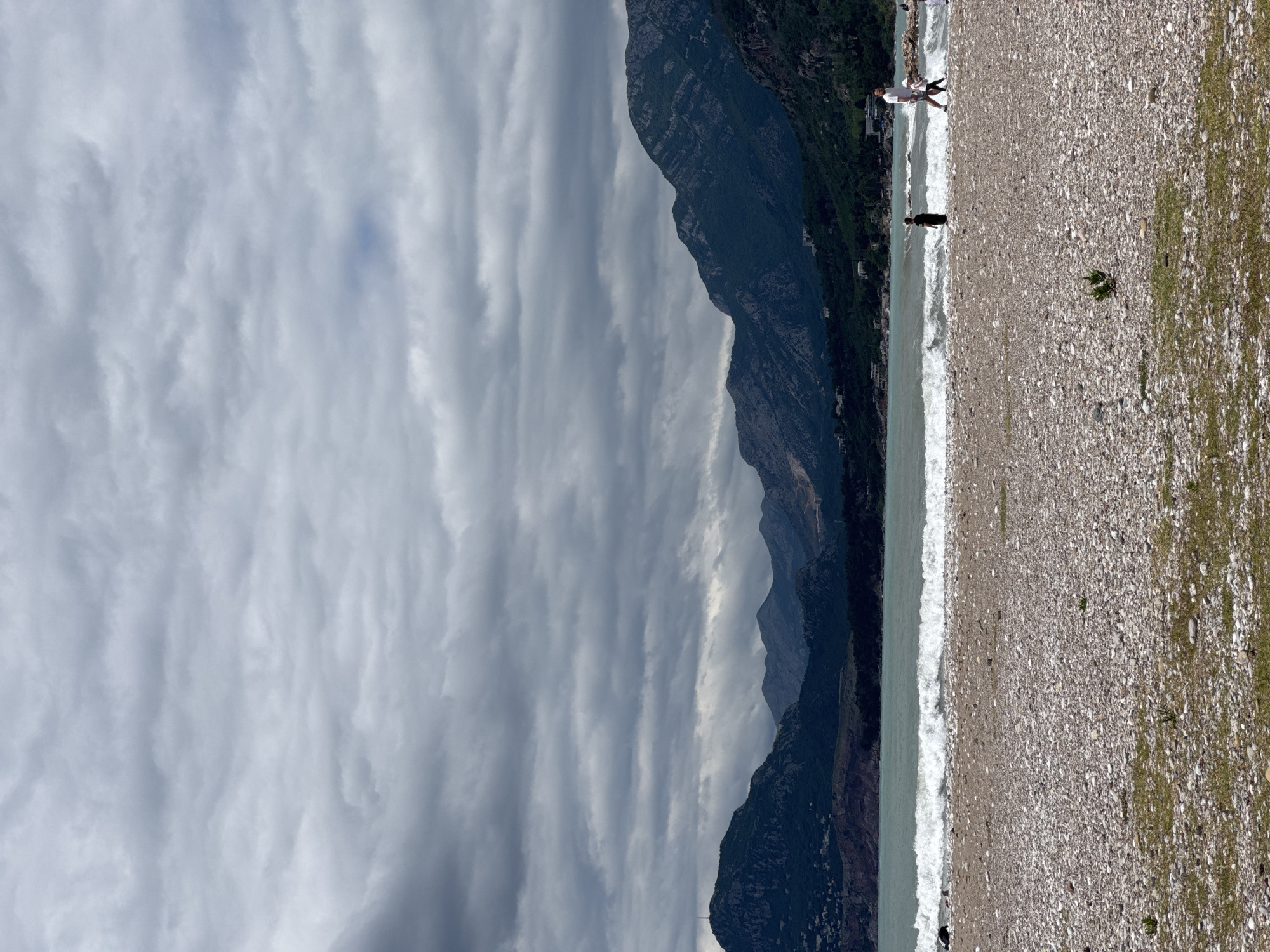The scene depicts a pebble beach in Bar, Montenegro, with a mountain range in the background and a cloudy sky overhead. The beach occupies the right side of the image and extends along the shoreline where gentle waves are visible. Two individuals are present: one person in dark clothing stands near the water's edge, and a pair of people, one in a white shirt, are further along the beach. The mountain range extends vertically to the left of the beach, and the sky, filled with layered clouds, occupies the remaining space.